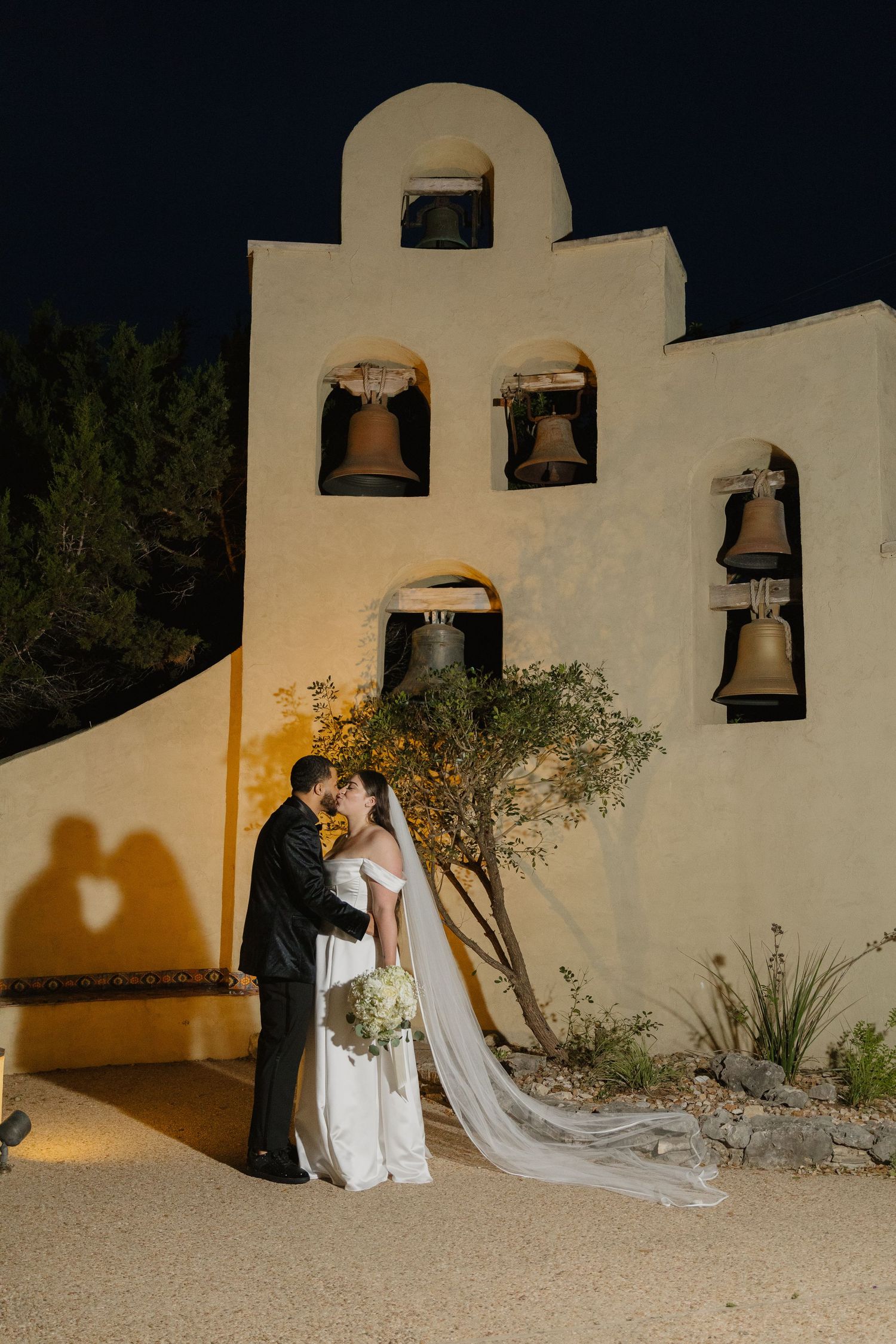 A couple shares a romantic moment at dusk in front of a Spanish mission-style bell tower illuminated by warm lighting.