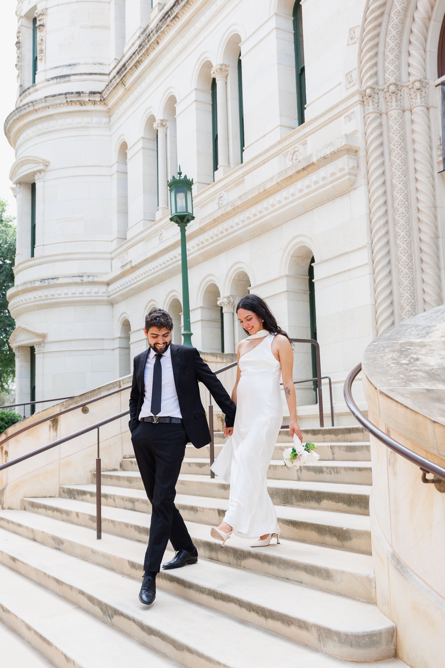 Bride and groom walking down grand stone steps of elegant building, she holds white bouquet in sleek gown, he wears black suit.