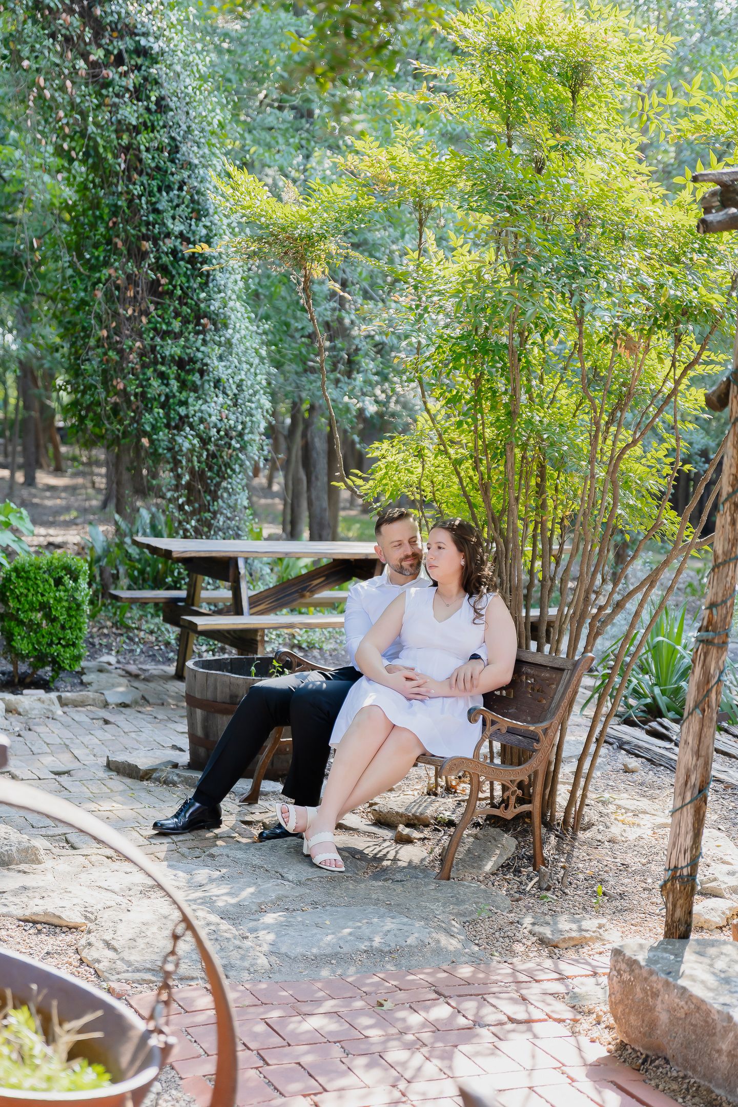 A couple relaxes on a wooden bench surrounded by lush greenery and bamboo in a peaceful garden setting.