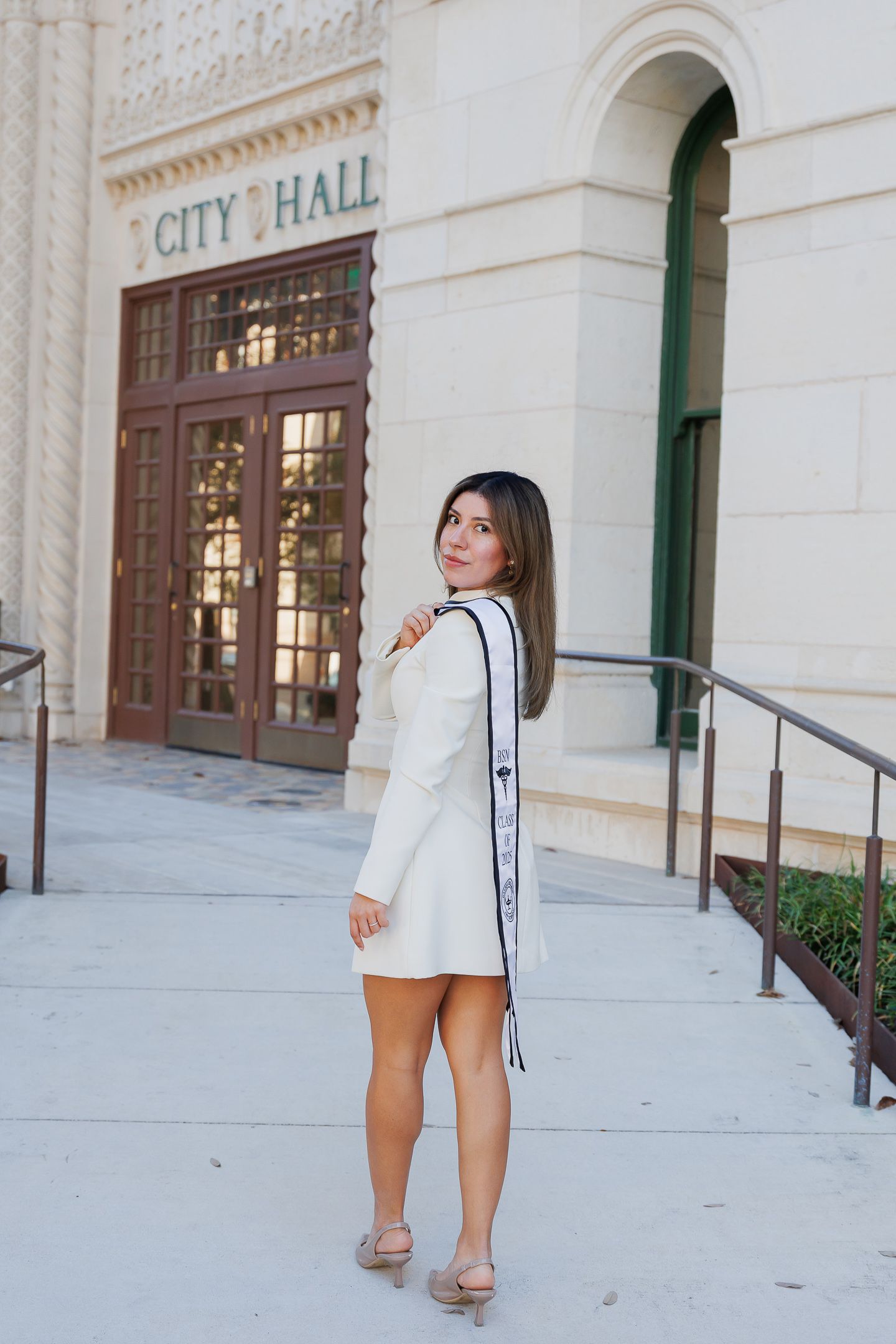 A person poses in front of City Hall wearing a white outfit and holding graduation stoles.
