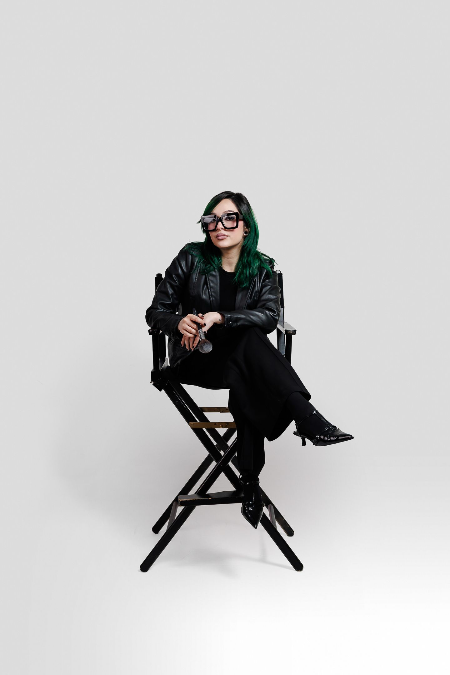 Person with green hair wearing black outfit and sunglasses poses in director's chair against white background.