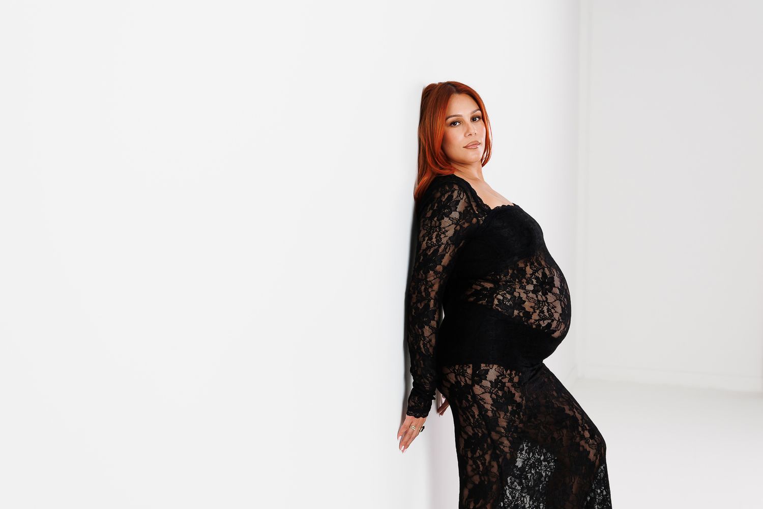 Pregnant woman in black lace maternity dress poses elegantly against white background.