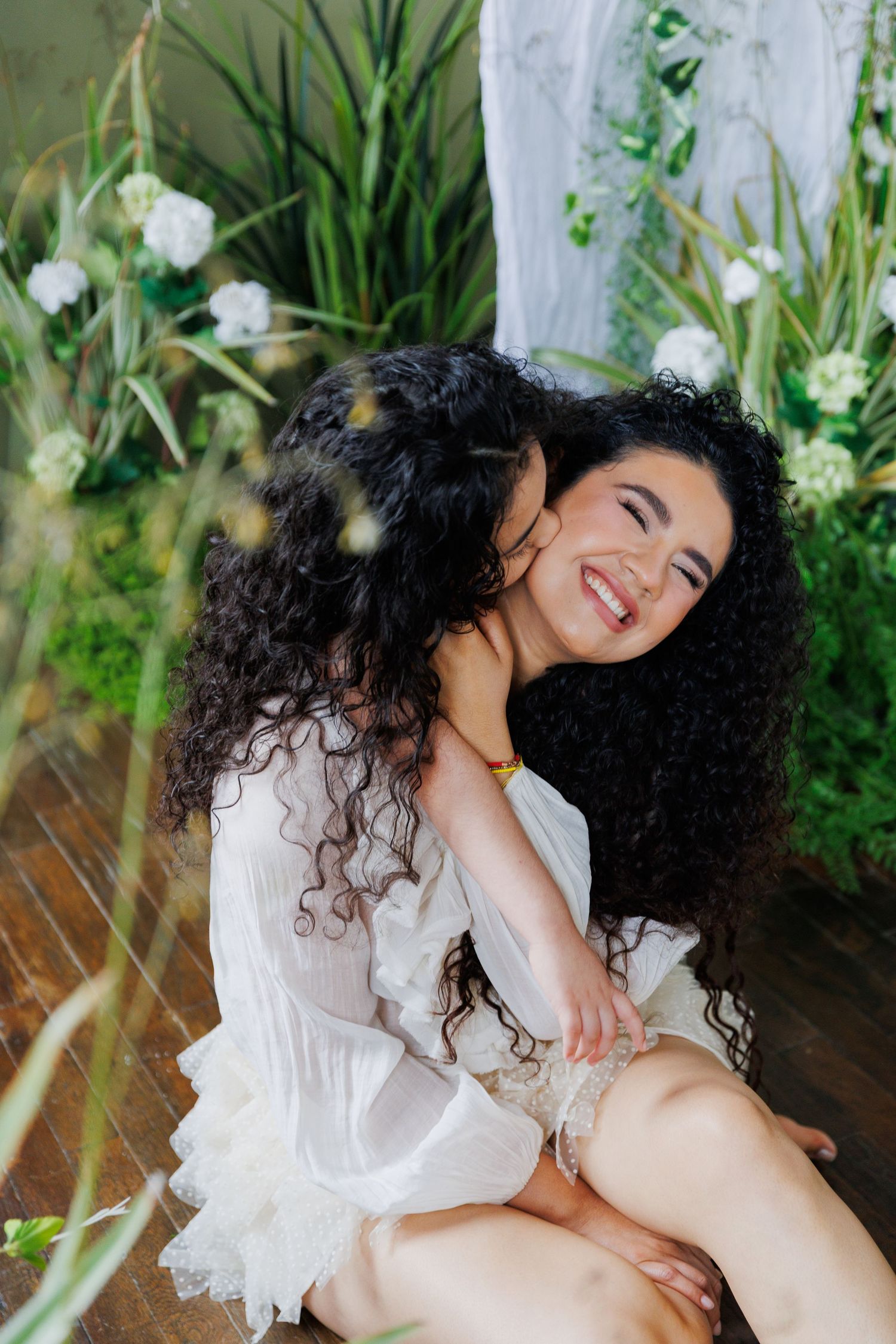 A person in a white dress sits outdoors surrounded by flowers and greenery, smiling with curly dark hair.