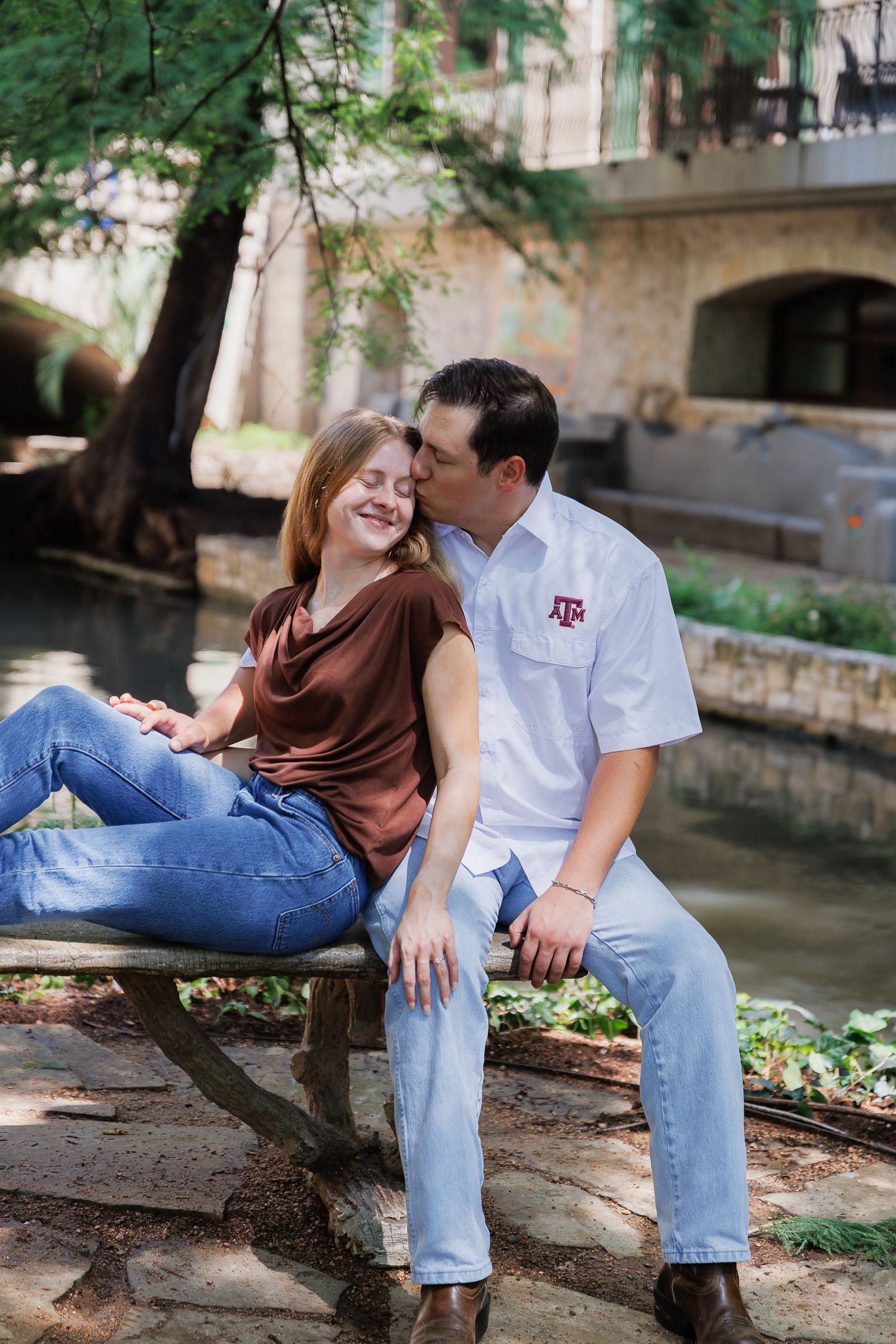 Couple sitting on bench by river, man kissing woman on cheek wearing Texas A&M shirt.