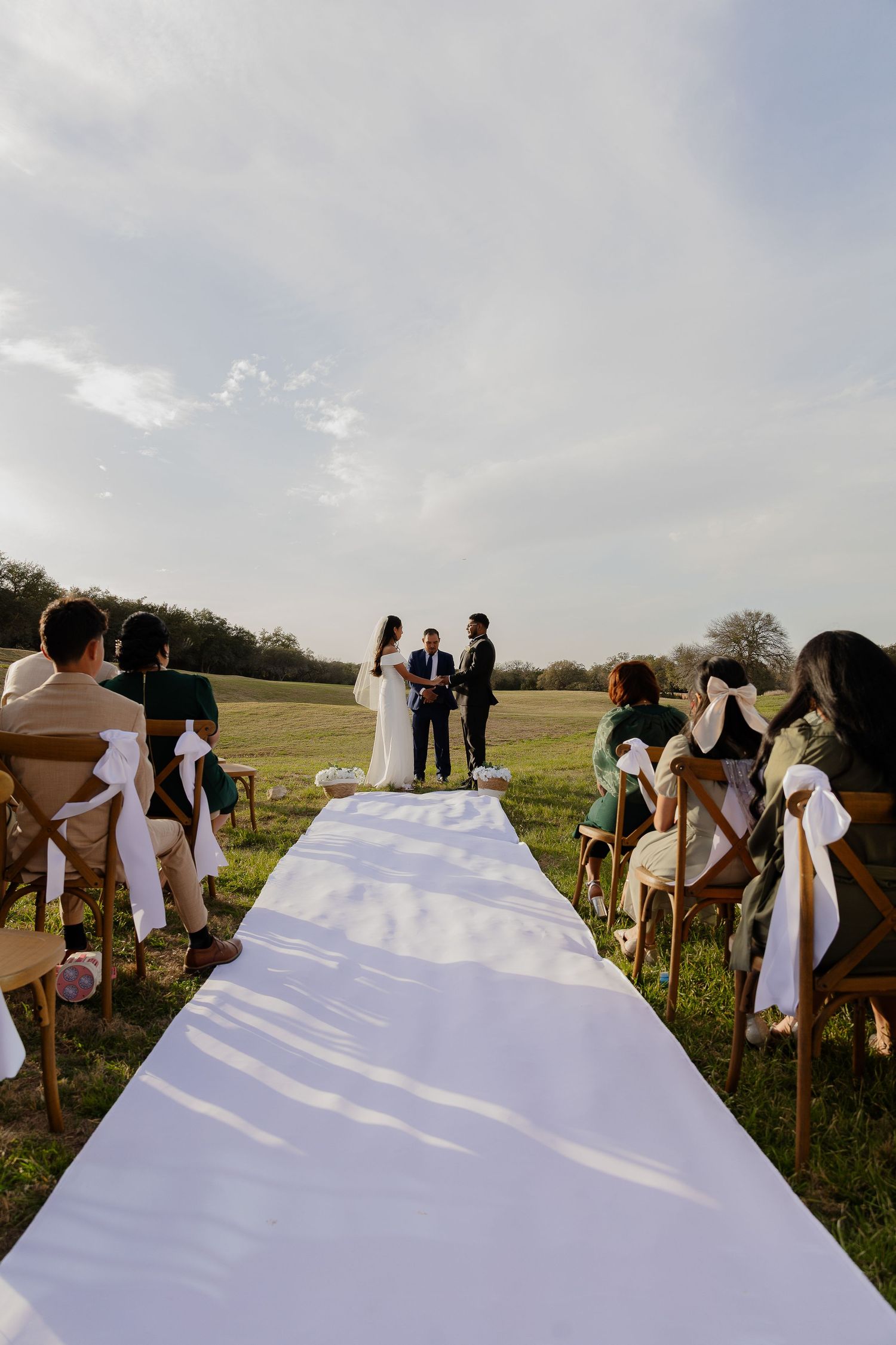 Wedding ceremony setup with white aisle runner and guests seated on lawn chairs under open sky.