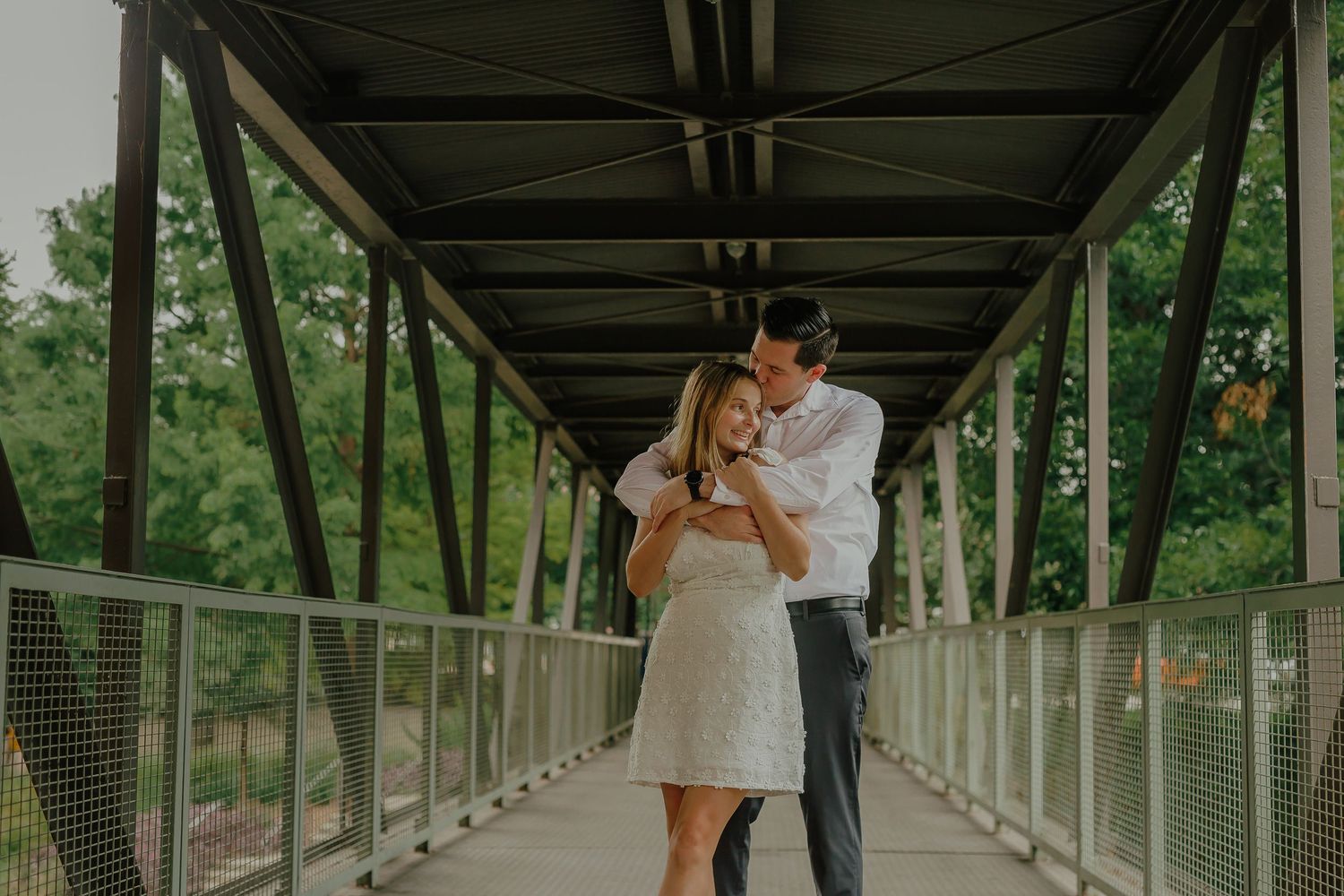 A couple shares an intimate moment on a covered bridge walkway surrounded by greenery.