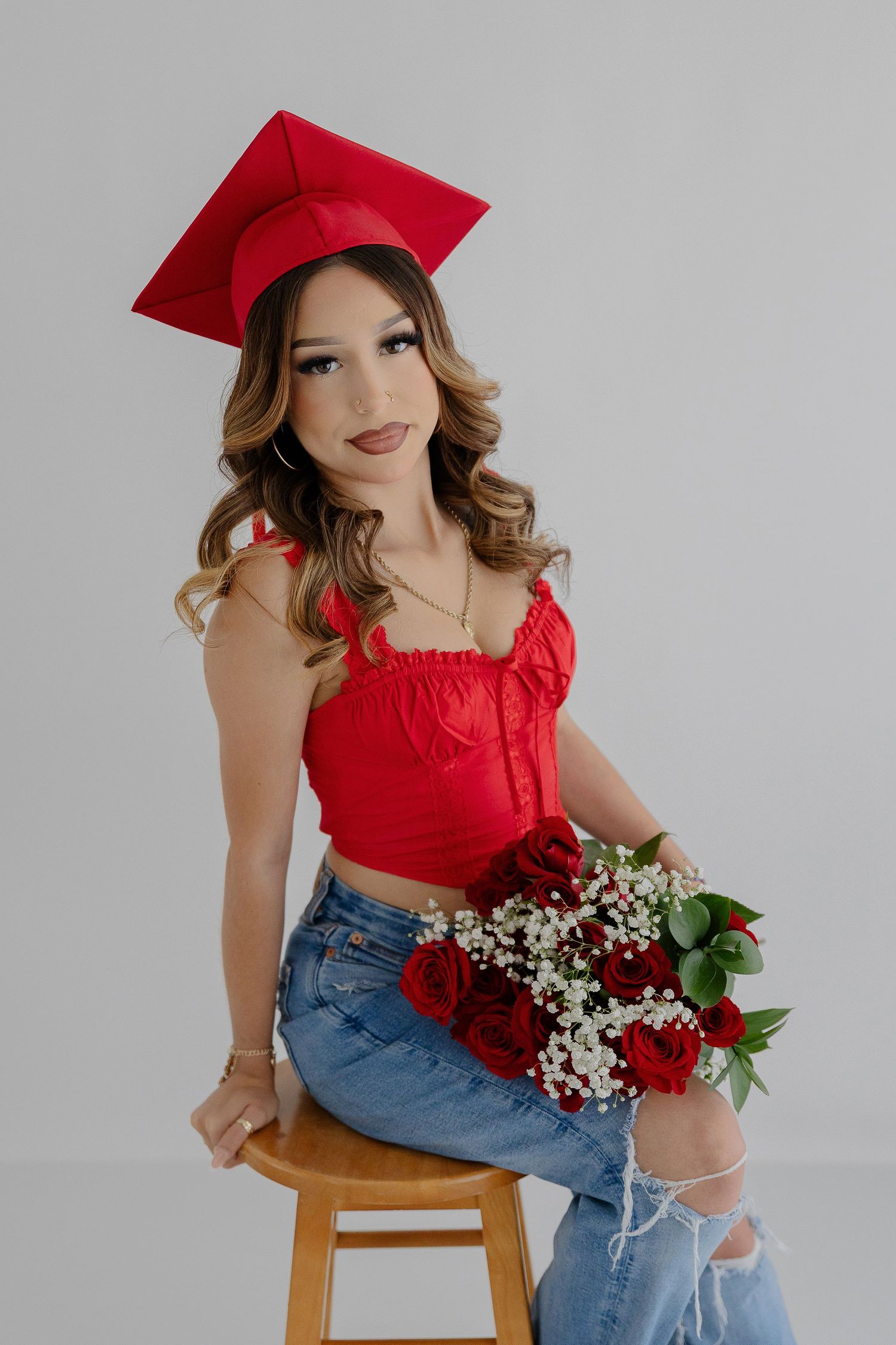 Graduate in red cap and casual attire sits on wooden stool with rose bouquet.