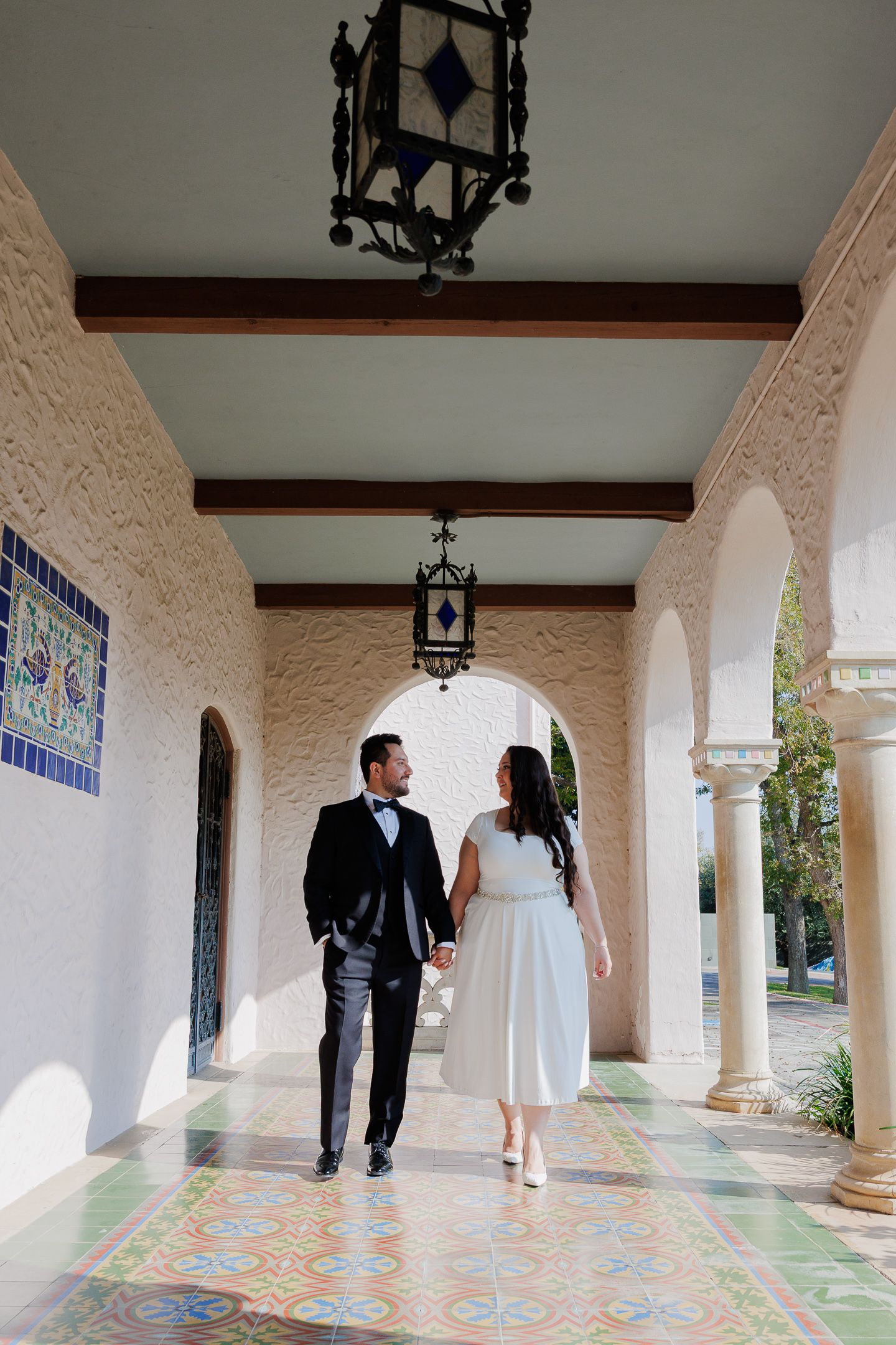 A couple walks hand in hand along an elegant Spanish-style colonnade with decorative tile floors and arched columns.