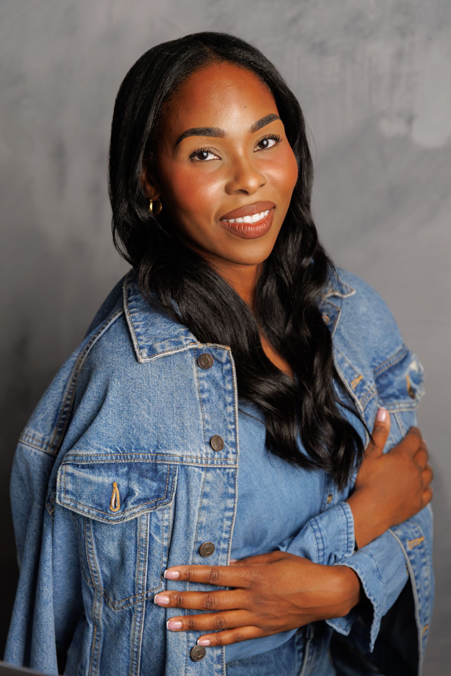 A smiling woman with long wavy black hair wearing a denim jacket poses confidently against a gray background.