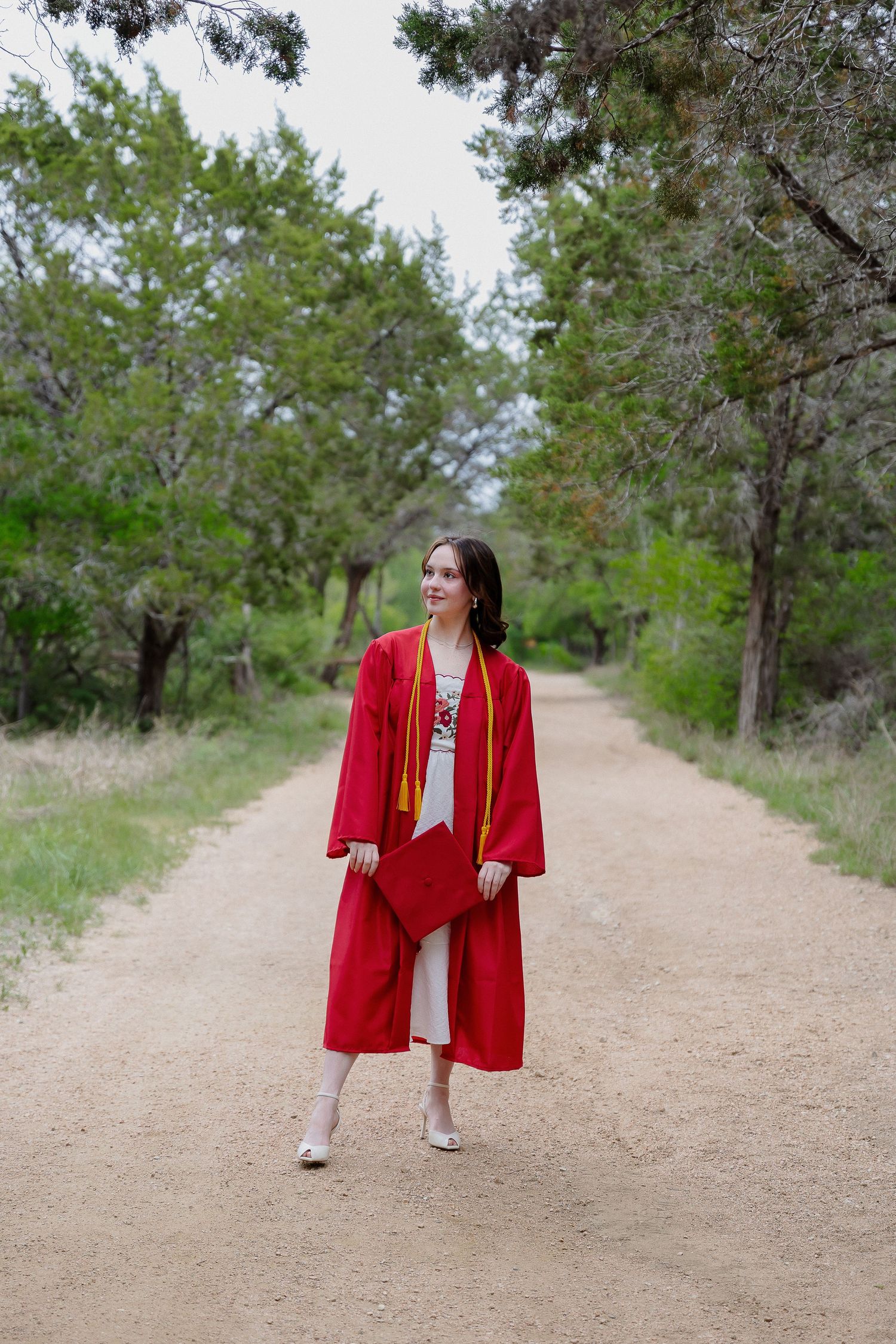Graduate in bright red academic regalia walking on rustic outdoor path.