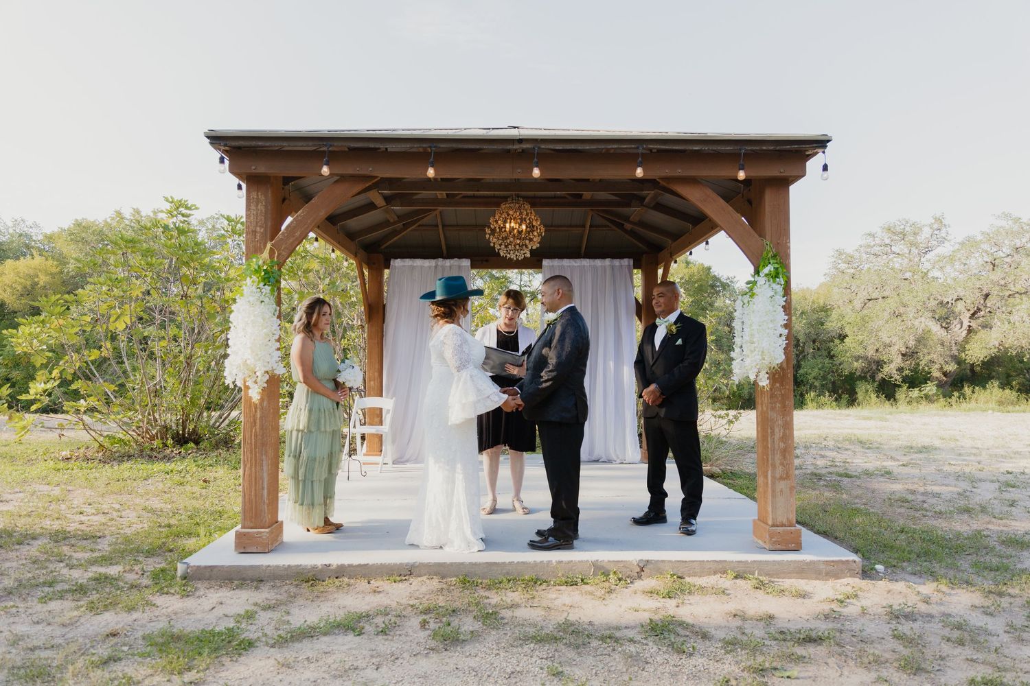Outdoor wedding ceremony under wooden pavilion with officiant and wedding party.