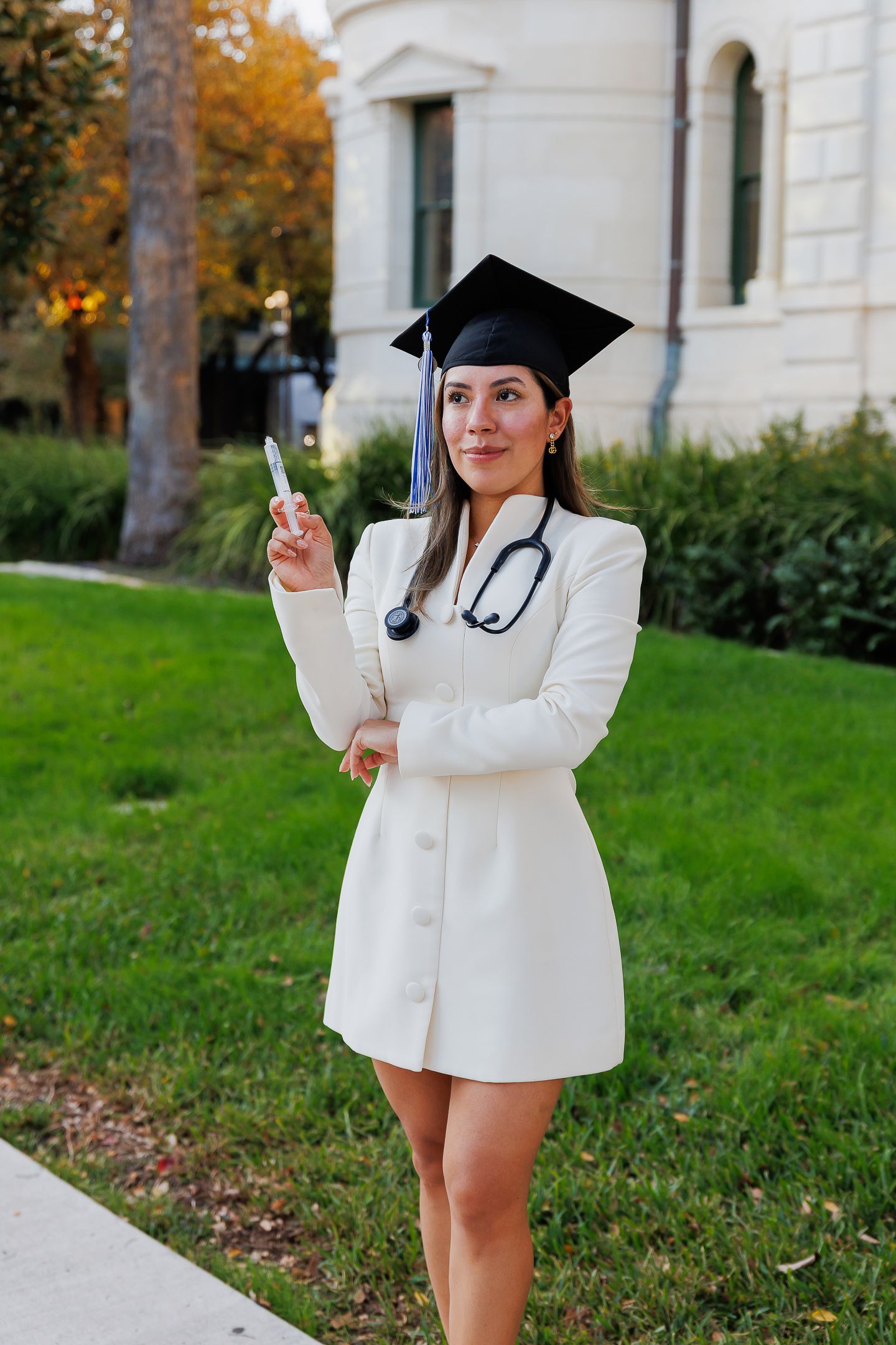 Graduate in white blazer poses outdoors during autumn with graduation cap and stethoscope against stone building.