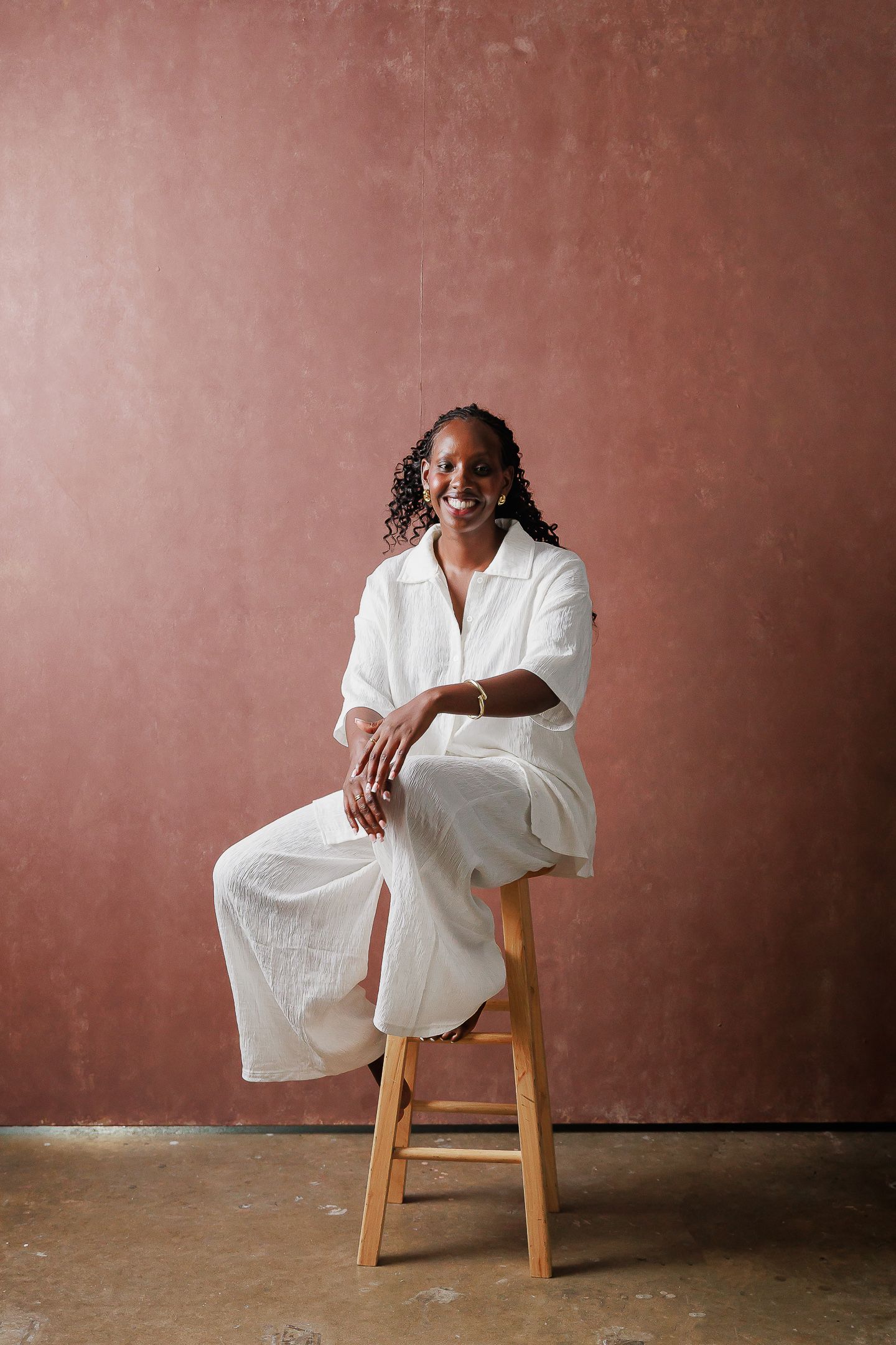 A figure in a flowing white dress sits gracefully on a wooden stool against a terracotta-colored wall in a minimalist setting.