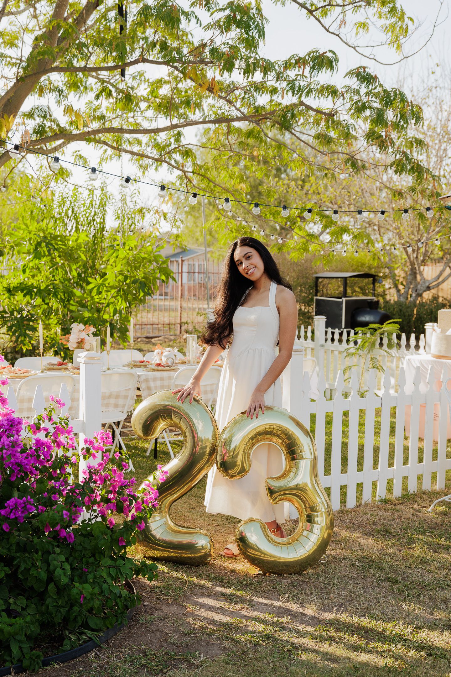Outdoor photo session in a garden with white picket fence, gold number balloons, and blooming pink bougainvillea flowers.