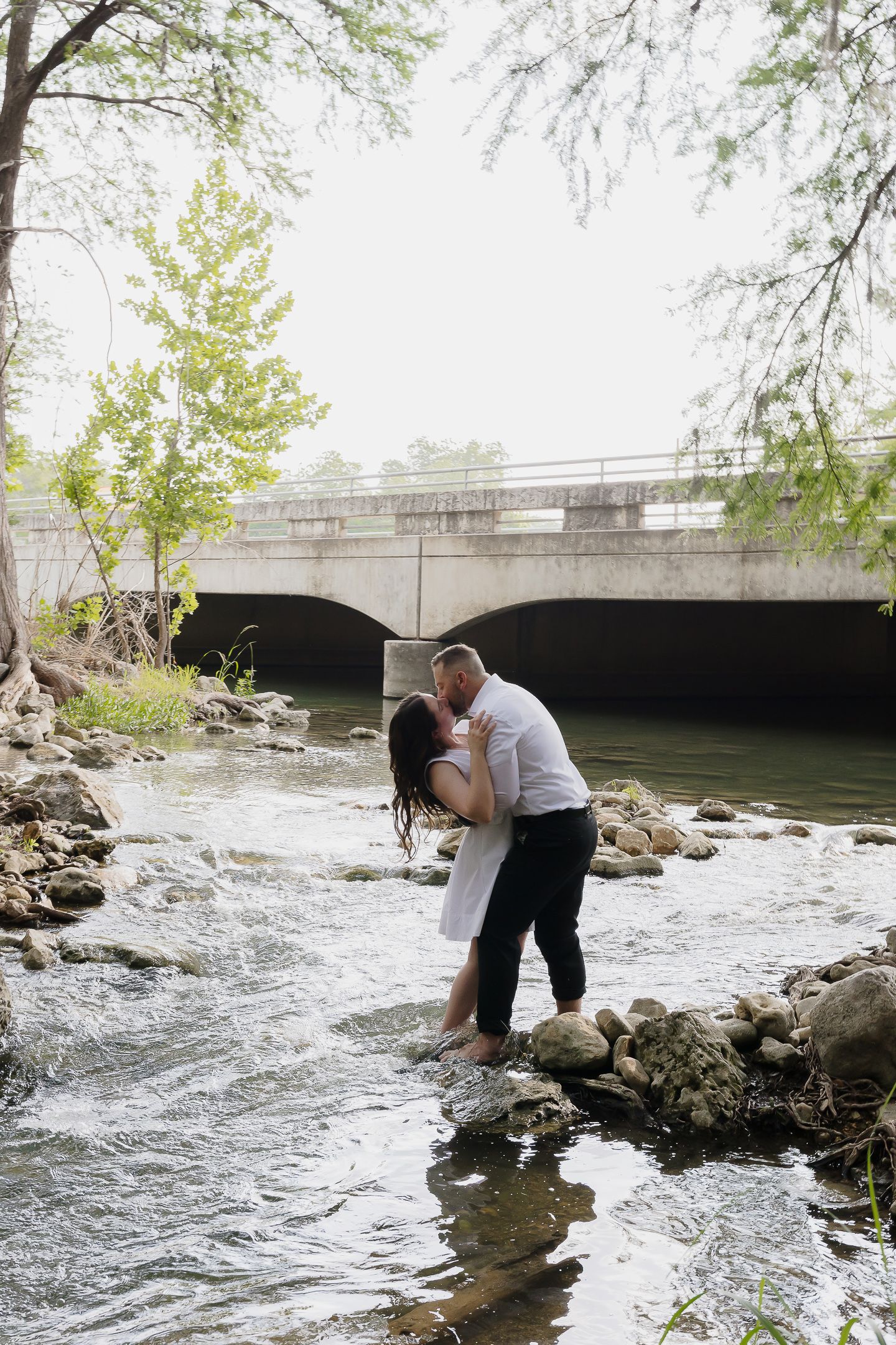 A couple shares a romantic moment by a rushing river under a stone bridge on a spring day.