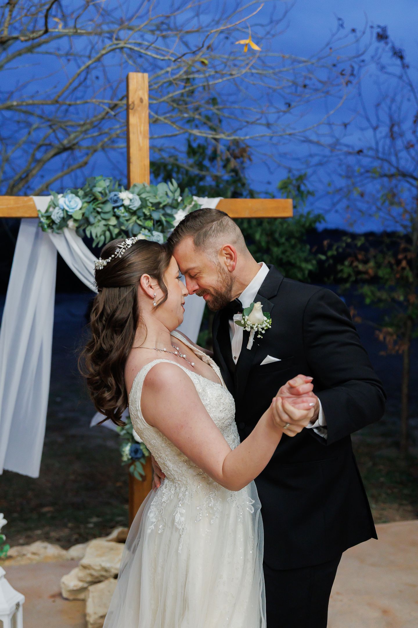 A romantic wedding couple shares their first dance outdoors under a wooden arch with white fabric draping.