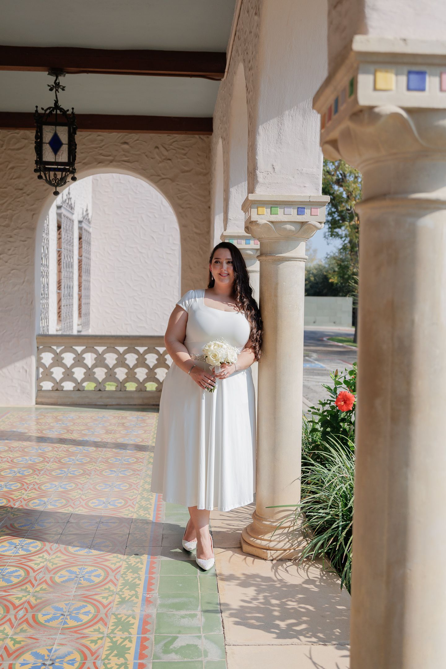 A series of white columns frame an outdoor walkway with decorative tiled flooring and greenery visible in the background.