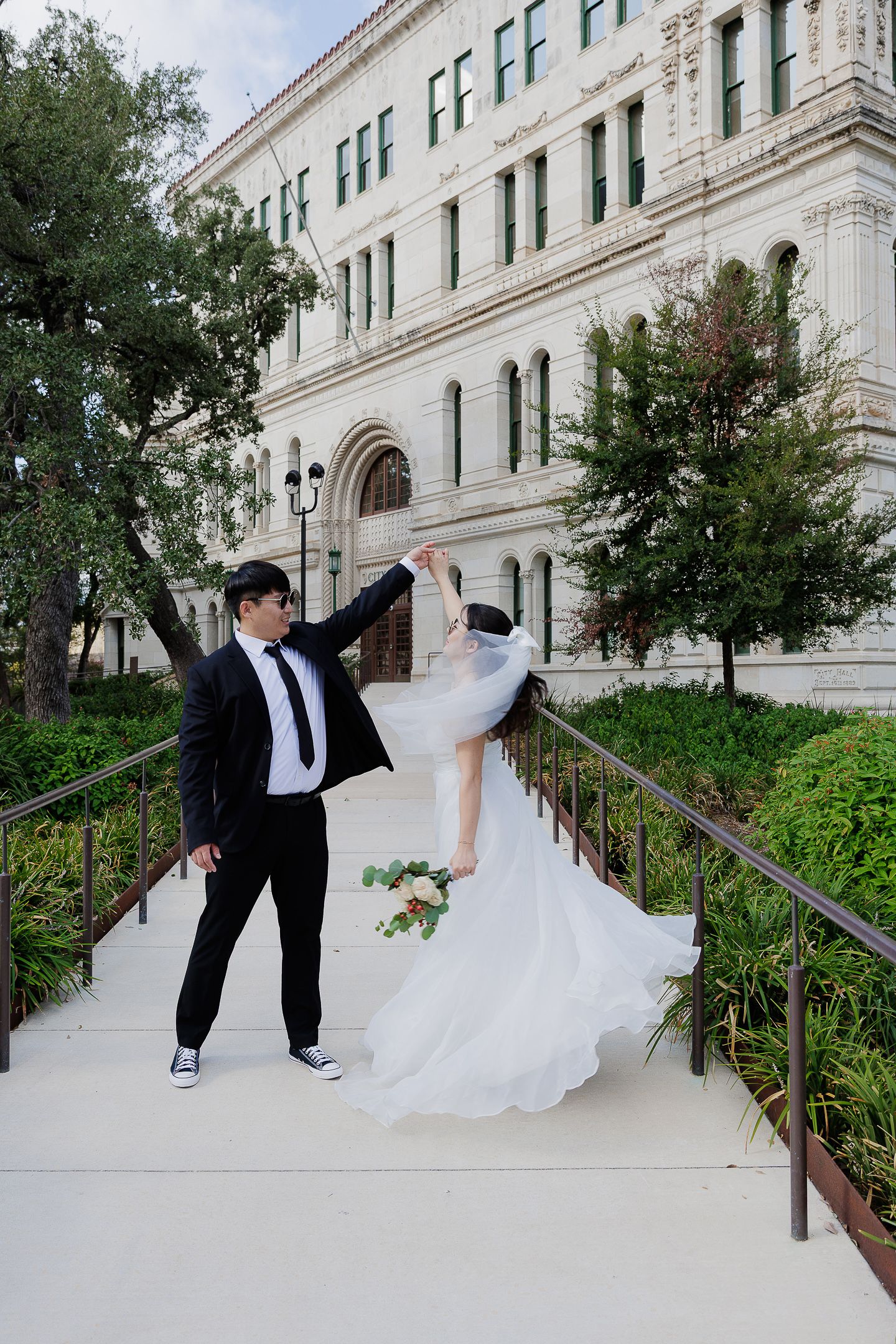 A couple in wedding attire poses playfully on the steps of a grand historic building with white stone architecture.