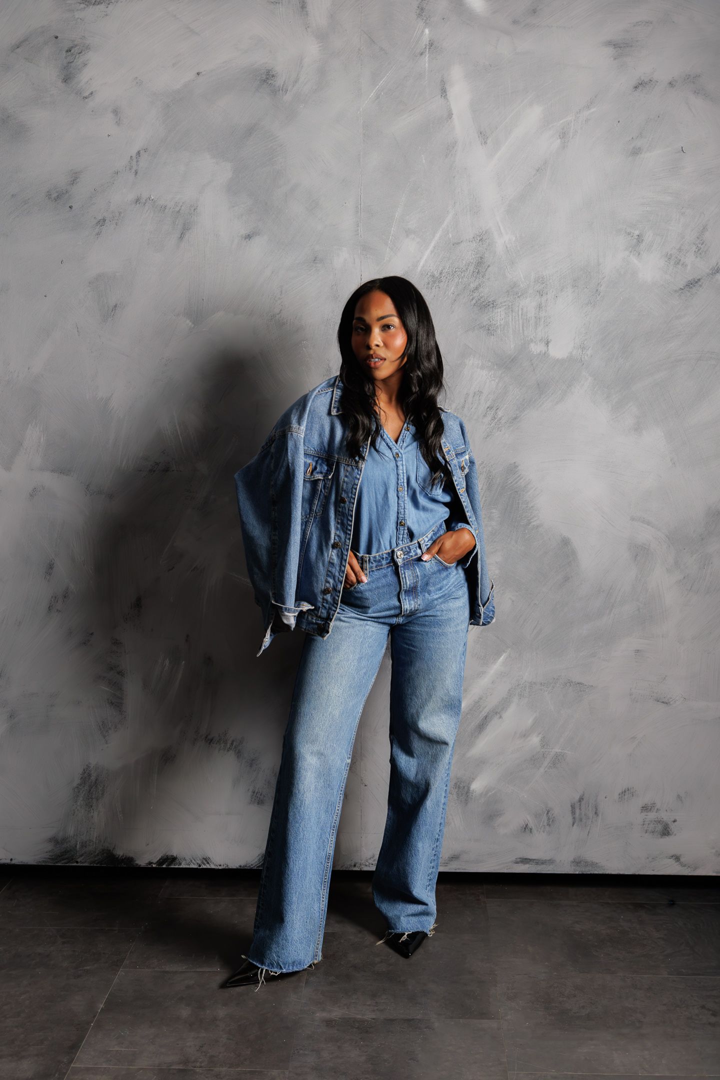 Woman in full denim outfit with wide-leg jeans and oversized jacket posing against gray textured wall.