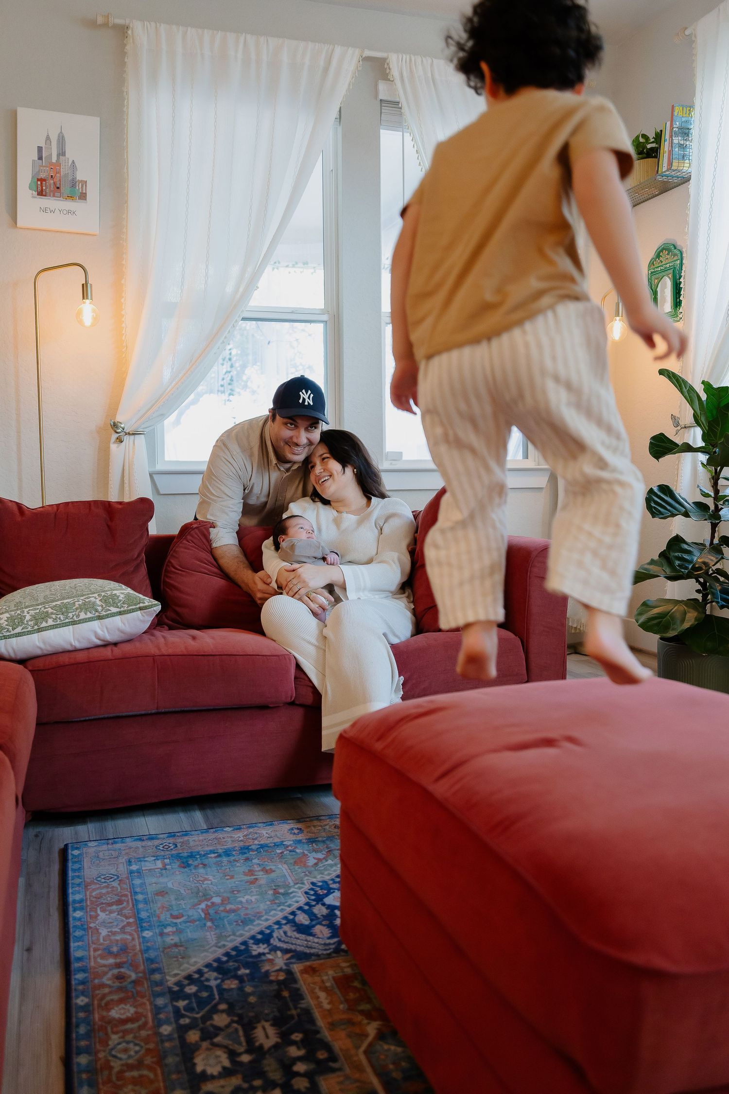 Family relaxing together in cozy living room with red couch, white curtains and decorative area rug.