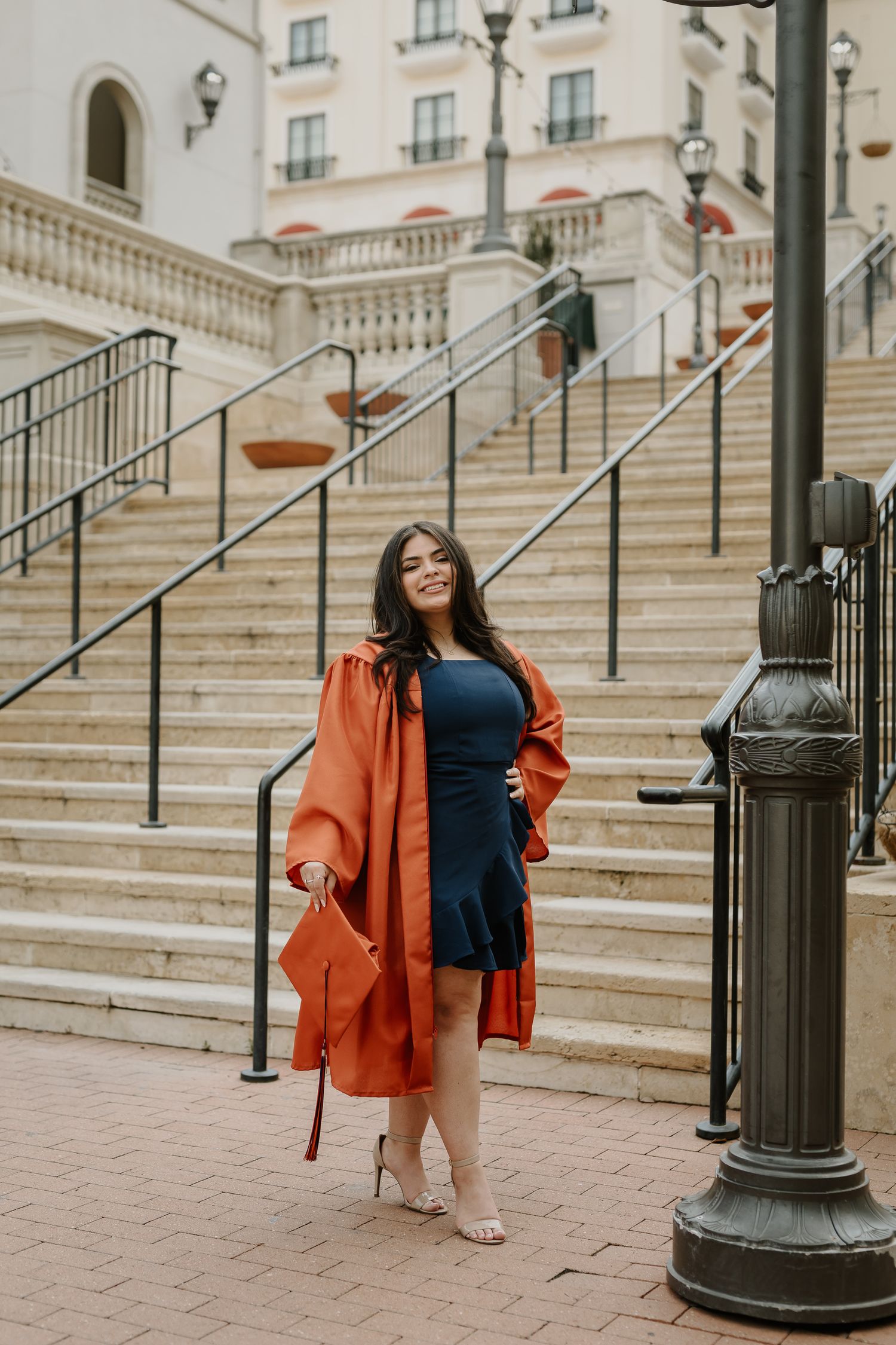 Graduate in orange coat poses on grand architectural steps during graduation photoshoot.