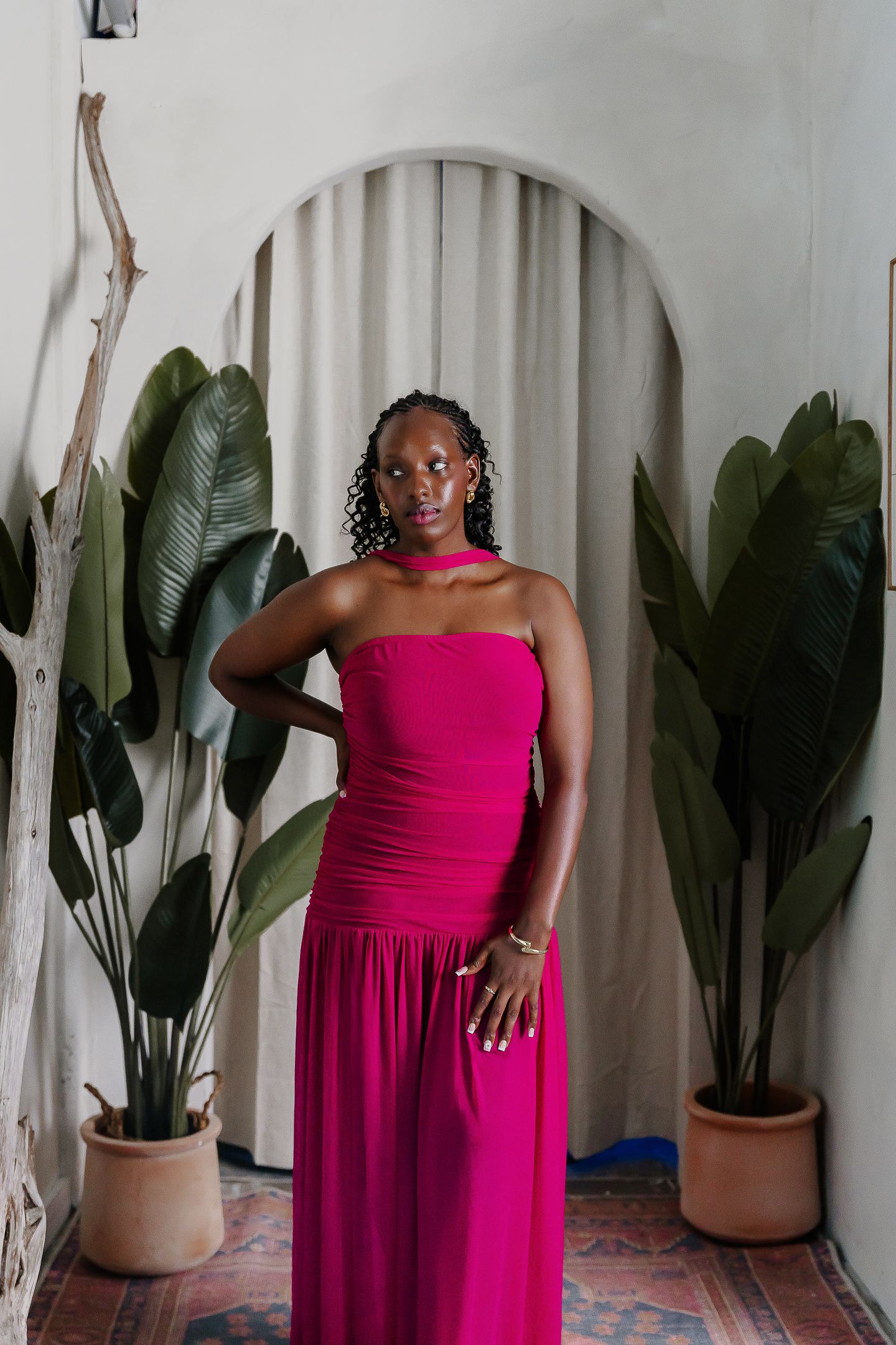 A woman in a stunning fuchsia evening gown poses elegantly against a white archway with potted tropical plants.