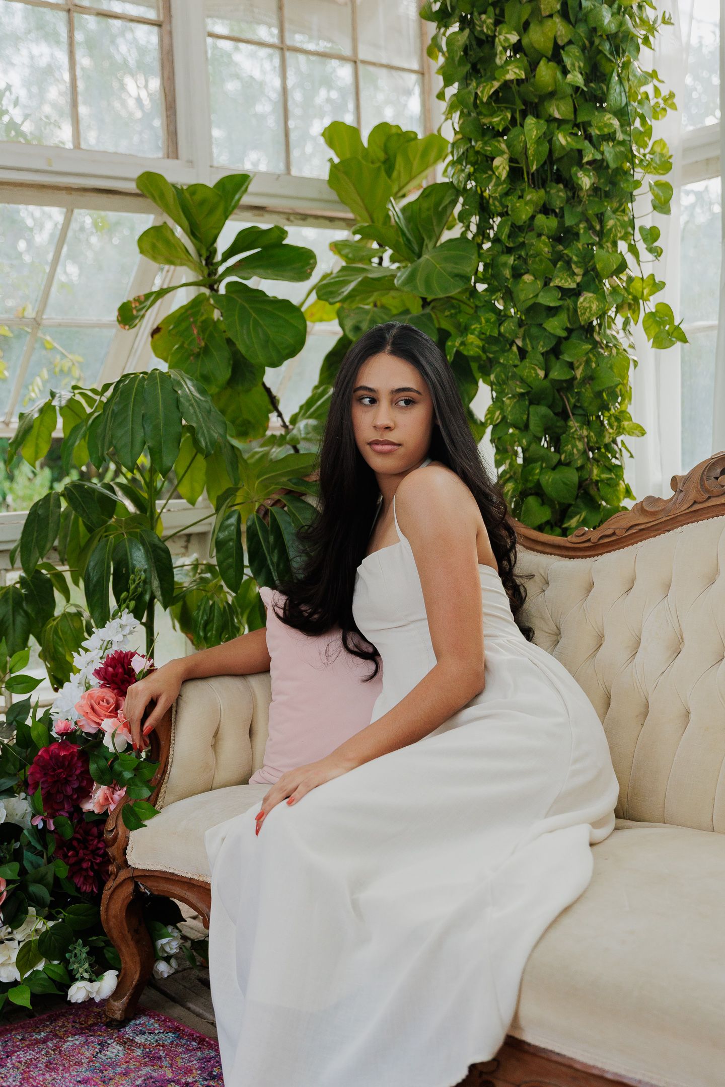 An elegant scene in a greenhouse with lush plants, vintage sofa, and flowing white dress.