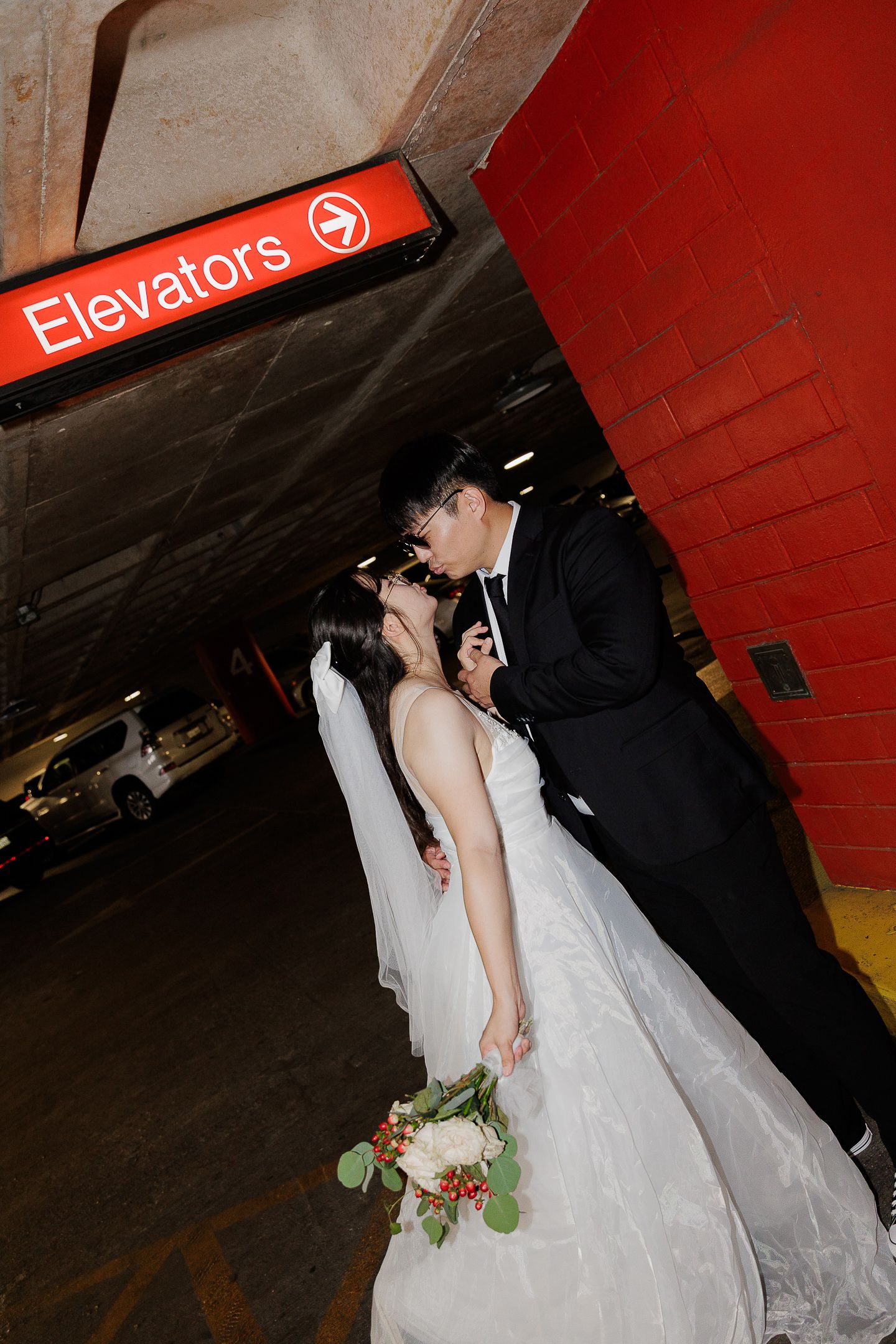 A couple in wedding attire share a romantic moment near an elevator sign in a subway station.
