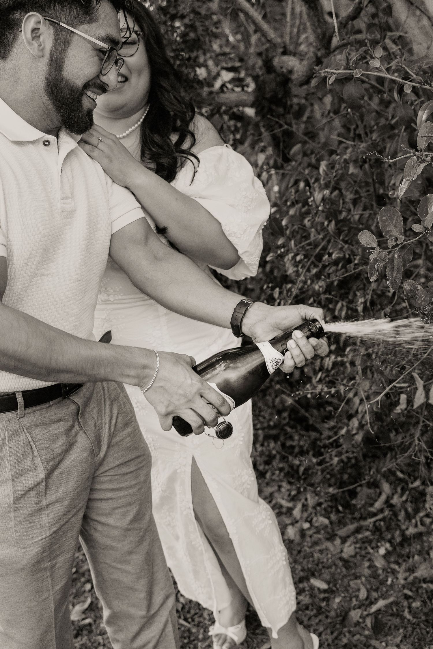 Black and white photo of couple popping champagne bottle outdoors.