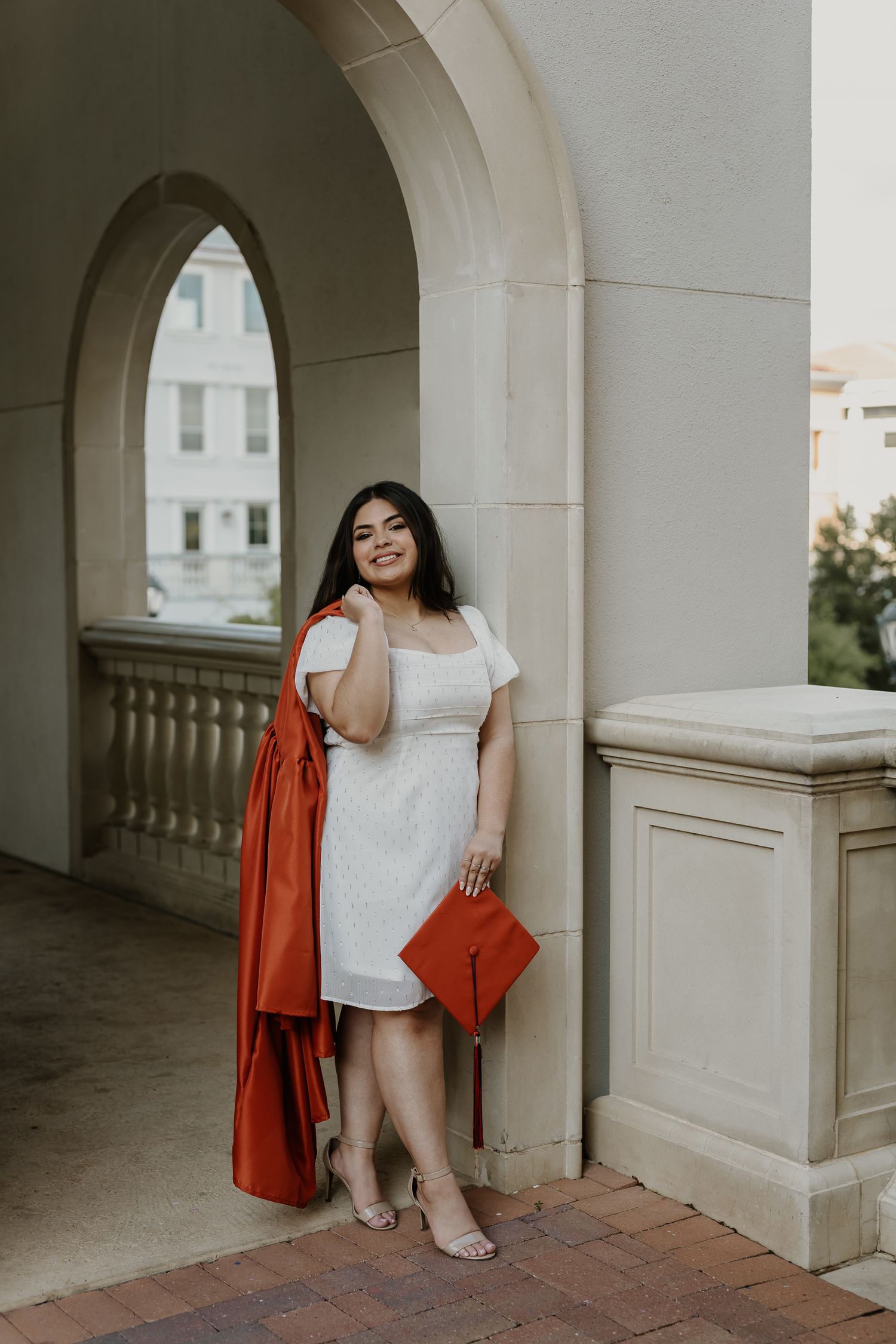 Graduate in white dress and orange graduation gown poses under elegant archway. San Antonio, Texas.