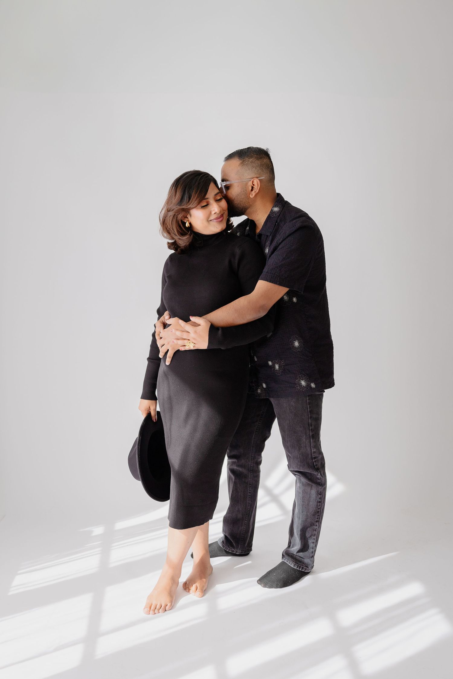 A couple in dark clothing shares an intimate embrace against a white wall casting dramatic shadows.