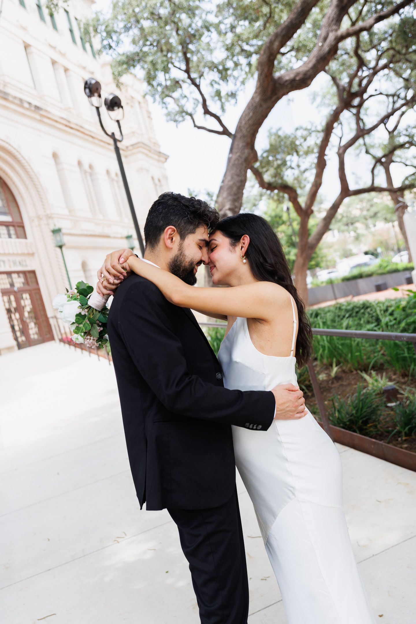 A bride and groom share a romantic moment outdoors, she in a white gown, he in a black suit holding a rose.