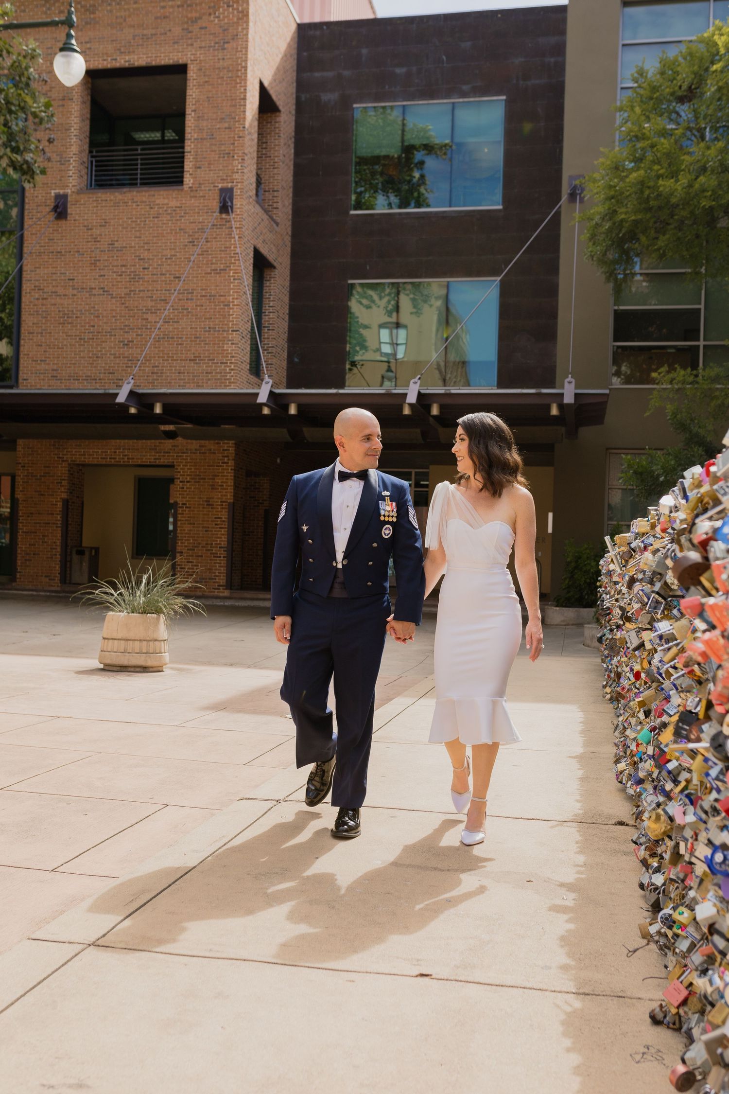 A couple walks hand in hand on an urban sidewalk with modern architecture in background.