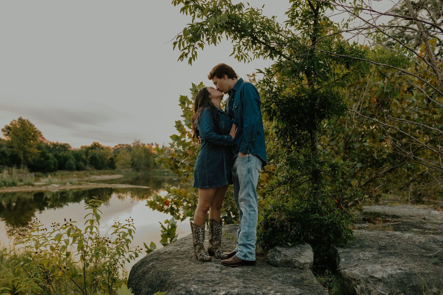 Couple shares intimate moment on rocky outcrop overlooking serene lake at sunset.