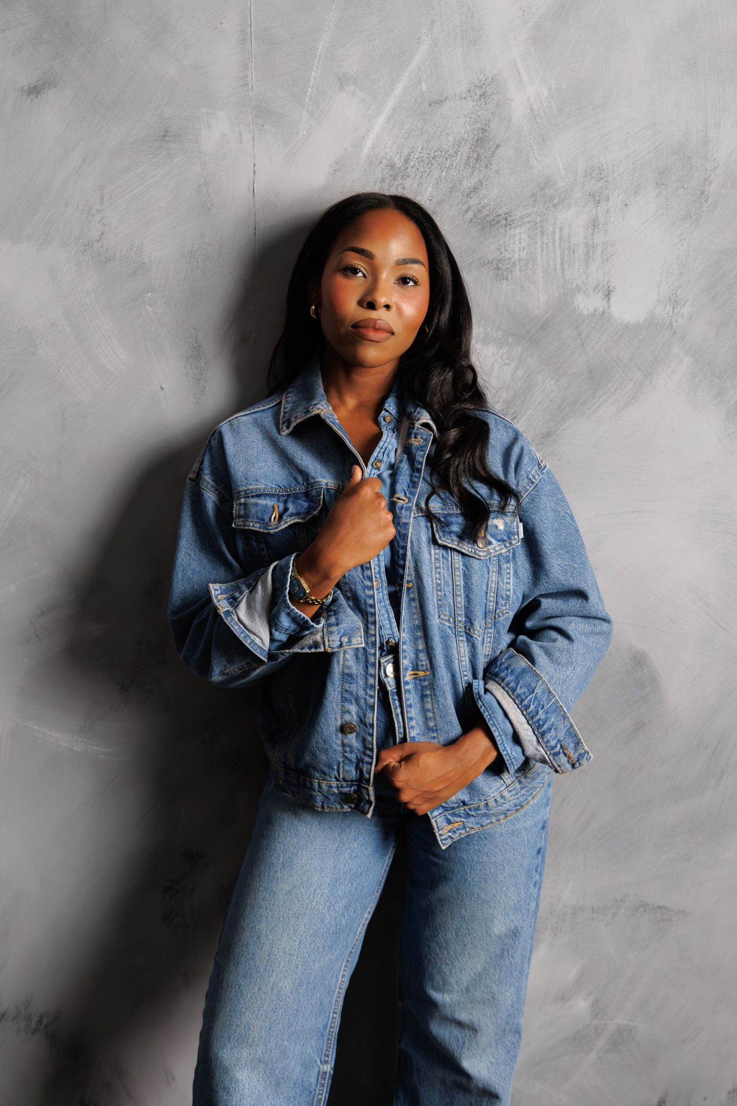 Young Black woman in denim jacket and jeans posing confidently against a gray textured wall.