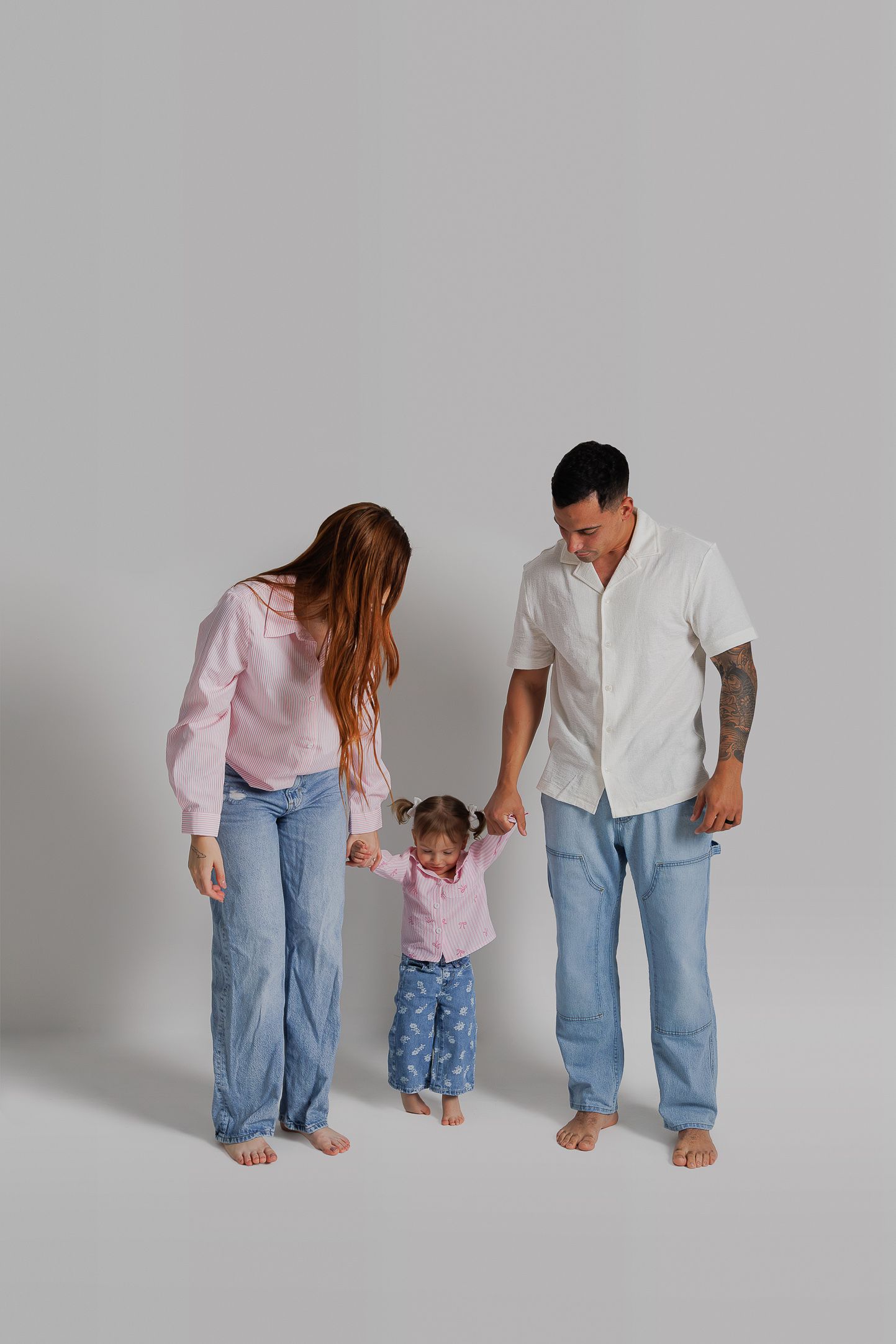 Family of three wearing coordinated casual outfits hold hands while standing barefoot against grey backdrop.