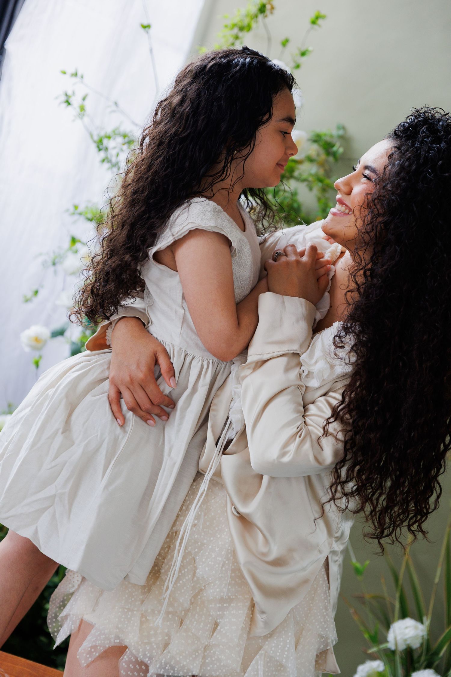 A tender moment between parent and child wearing matching cream-colored outfits against a light backdrop with greenery.