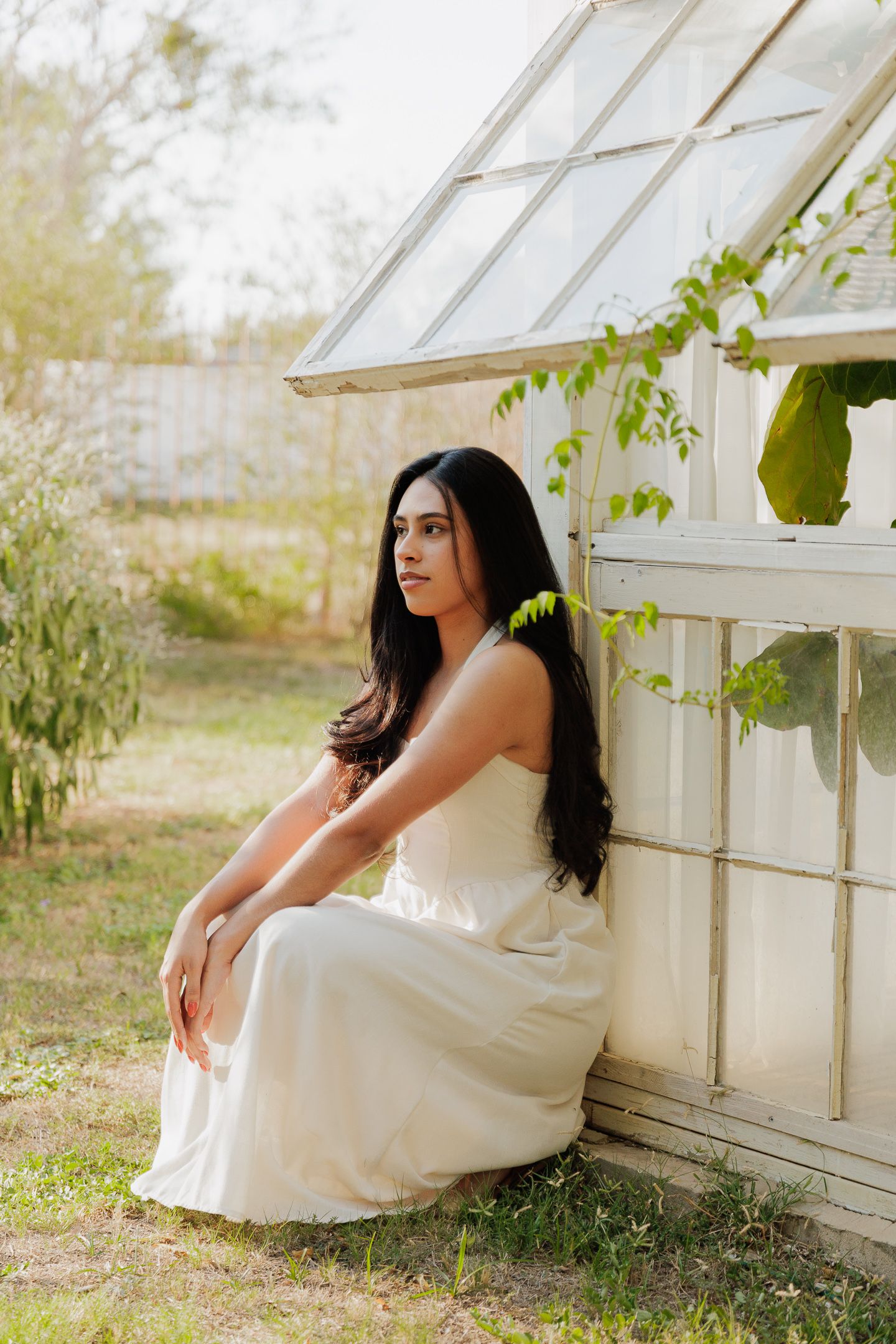A white dress-clad figure sits gracefully beside a sunlit greenhouse with climbing vines.