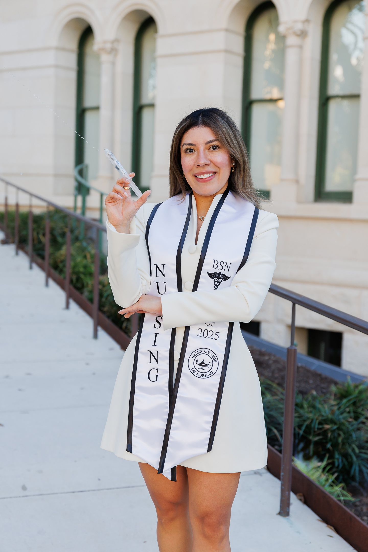 A graduate poses with decorated stoles and regalia outside a historic building with arched windows.