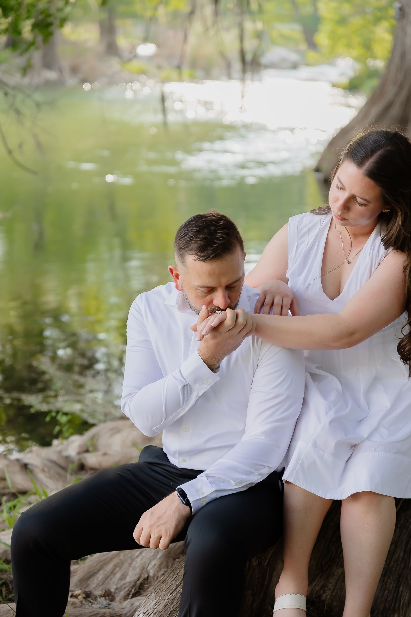 A couple in white clothing shares an intimate moment while sitting on rocks near a pond in a natural setting.