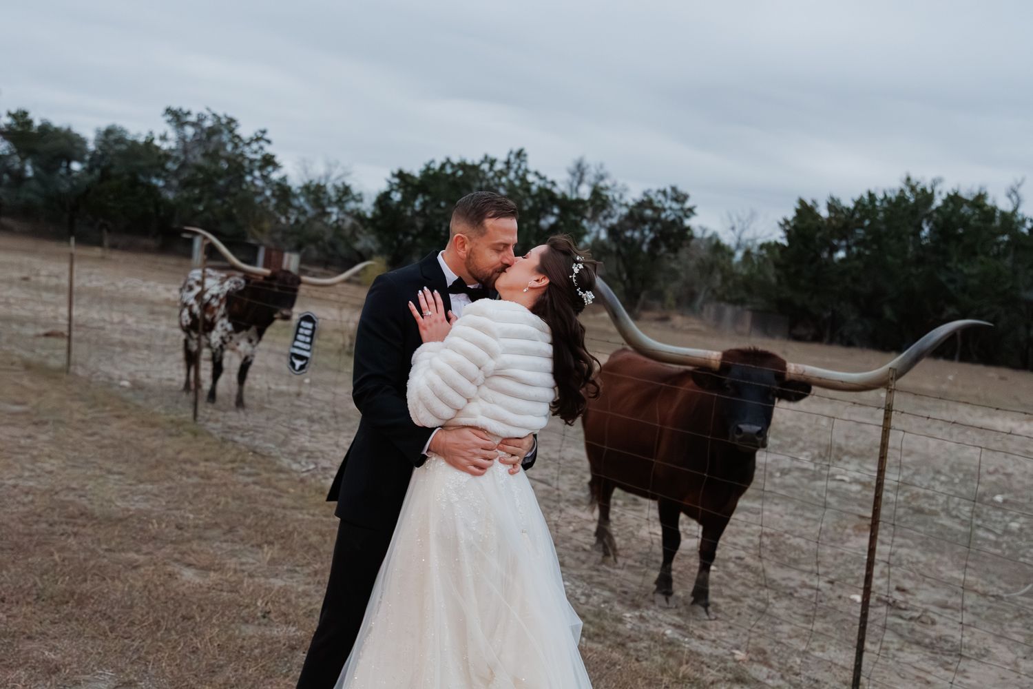 A couple shares a romantic moment outdoors with horses grazing in the background during a rustic countryside photoshoot.