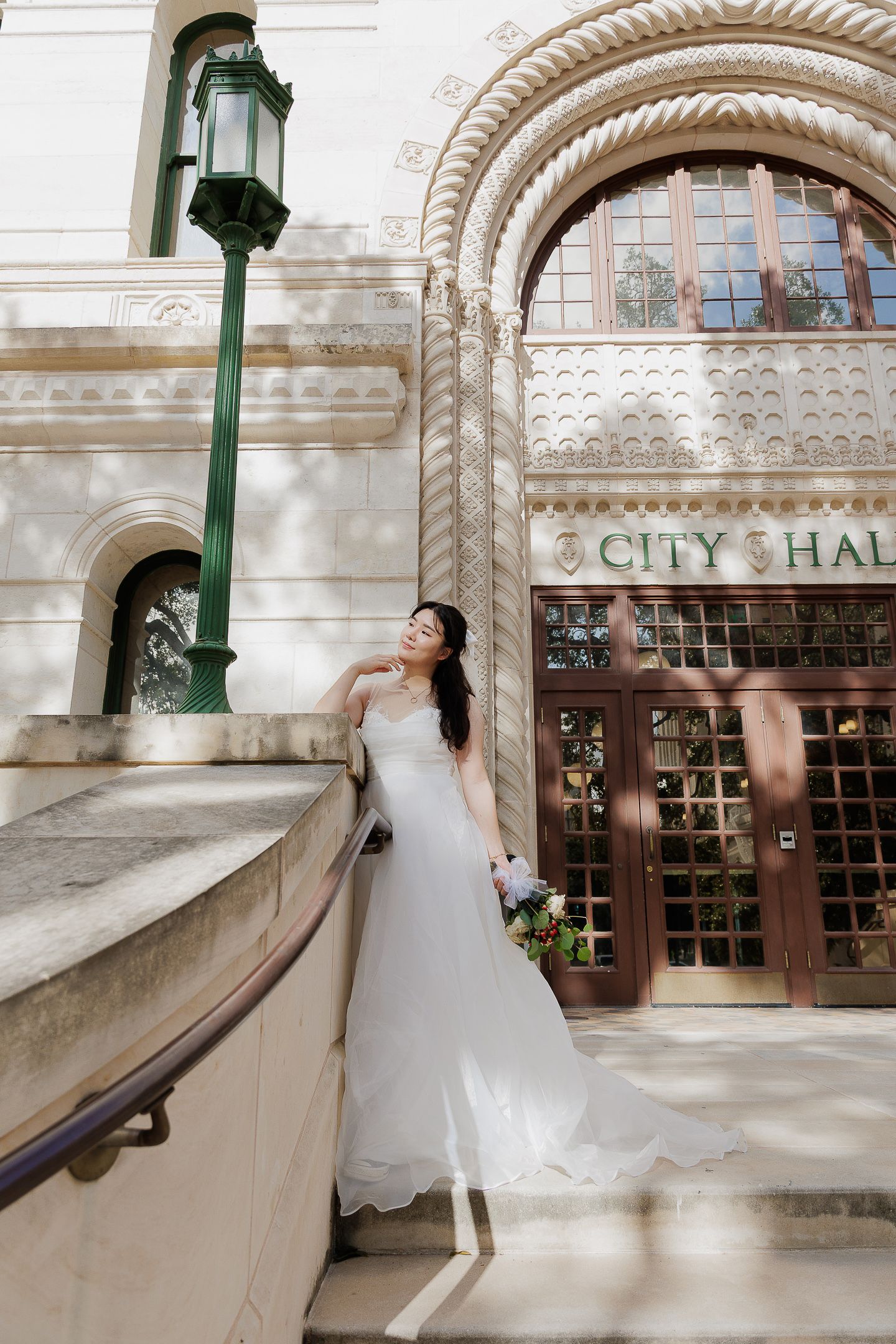 Bride in flowing white dress poses elegantly on City Hall steps with ornate architectural details visible.