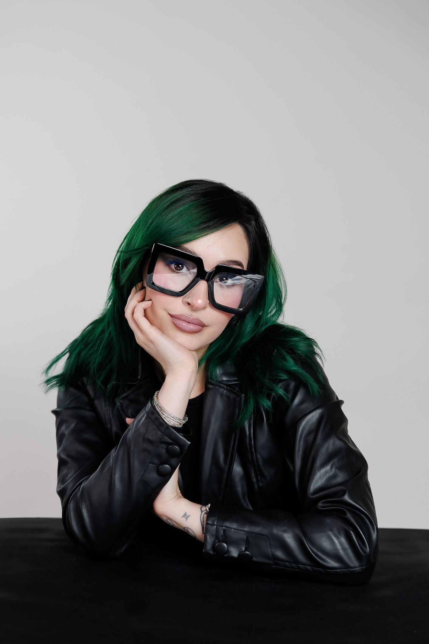 Person with green hair wearing glasses and black leather jacket poses against white background in stylish photo series.