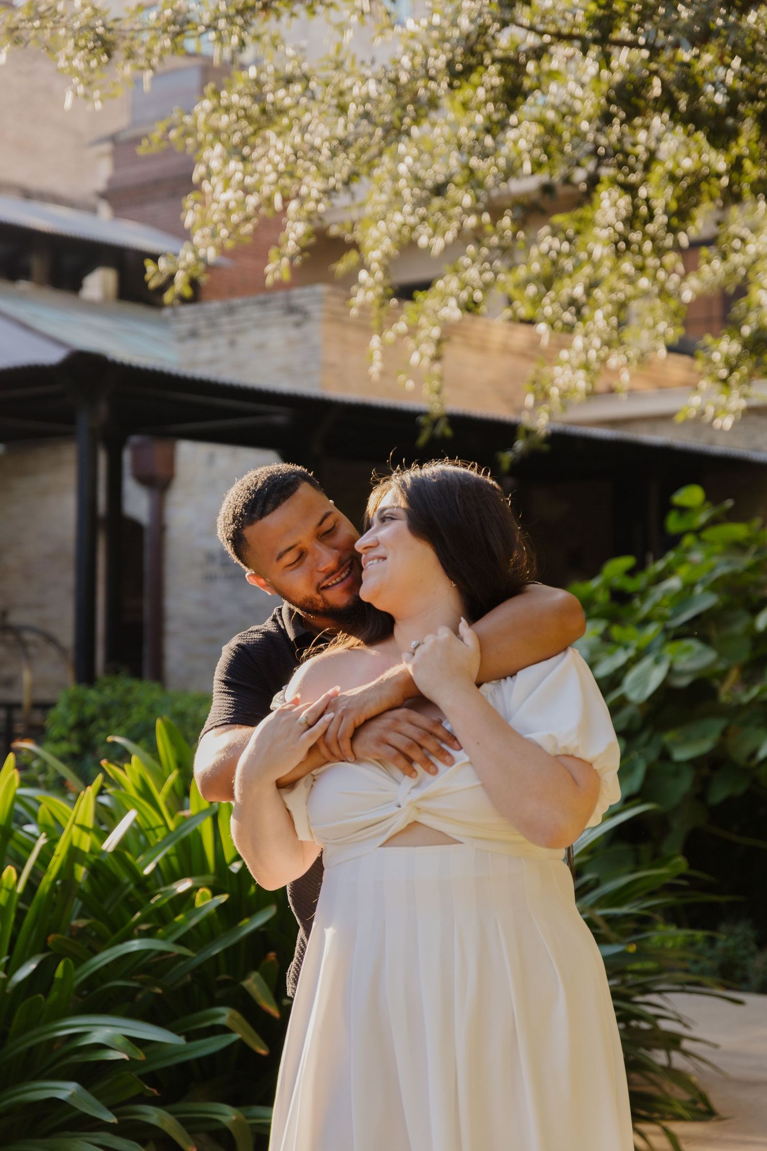 Couple embraces in a sun-drenched garden beneath flowering tree branches.