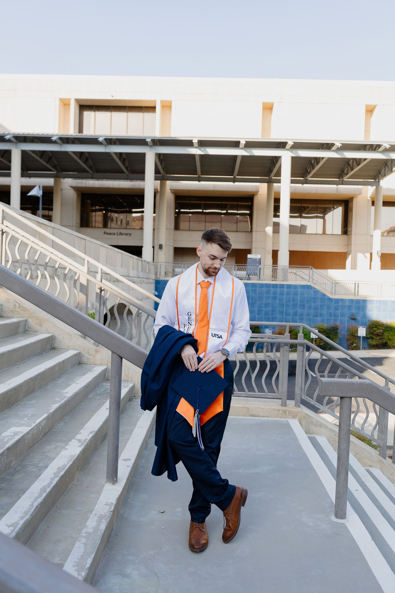 Graduate descends concrete stairs in navy graduation gown with orange stole.