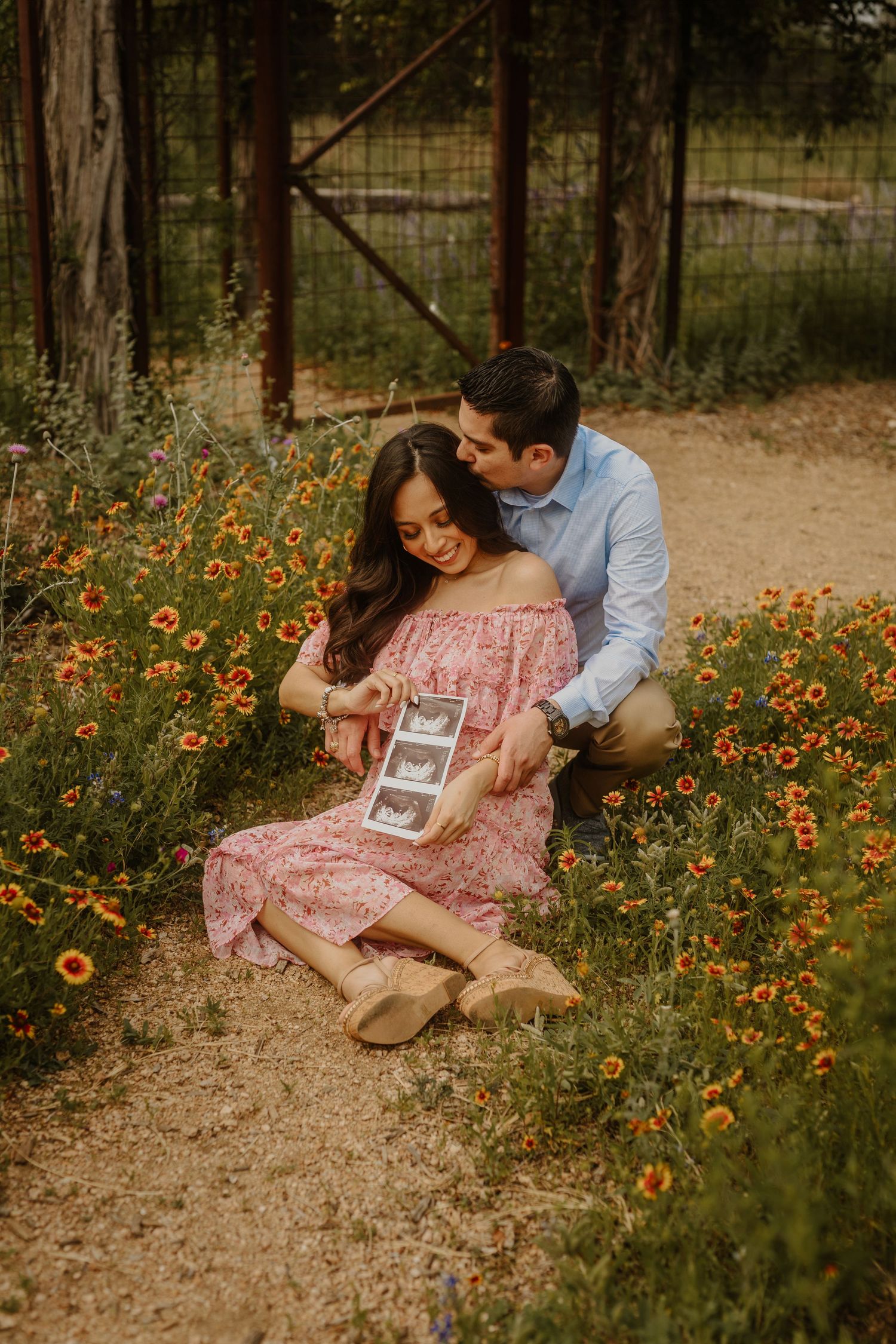 Couple shares intimate moment among vibrant wildflowers on dirt path as they hold ultrasound photos.
