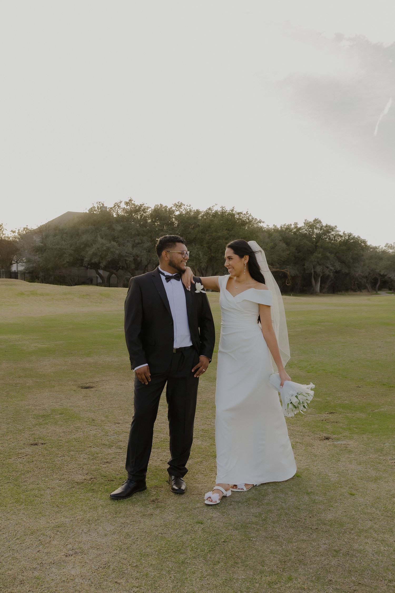 A wedding couple poses together on a grassy field during sunset.