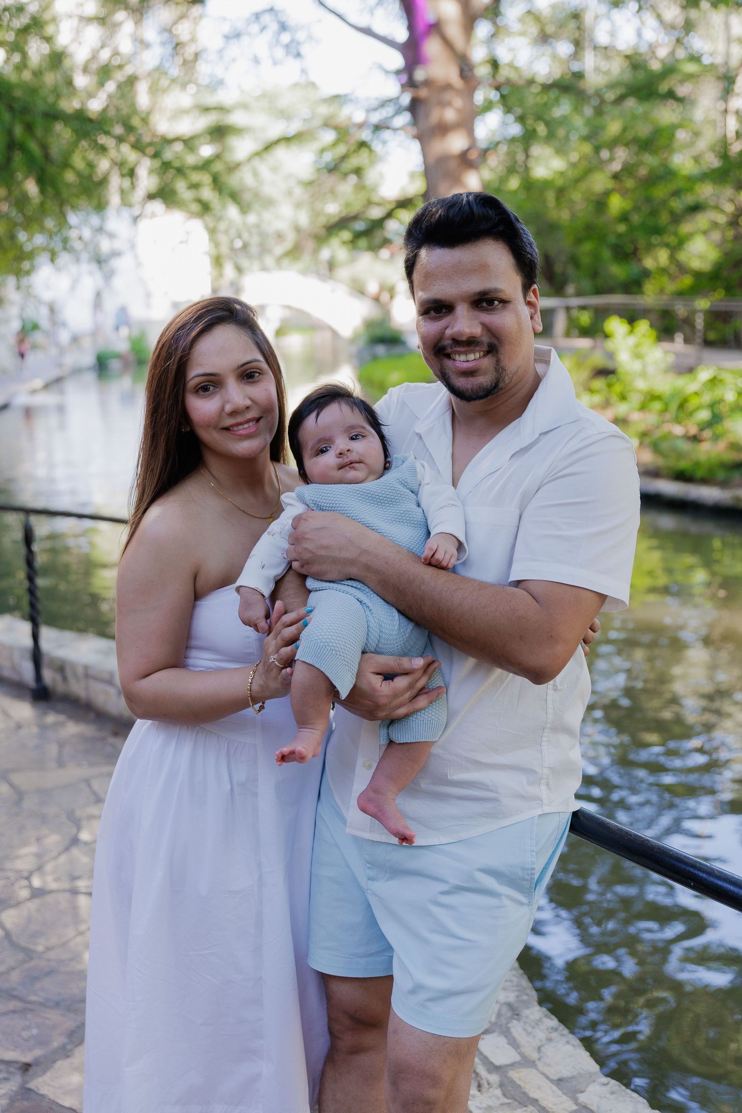 A couple in white outfits holds their baby near a waterfront during a family photoshoot.