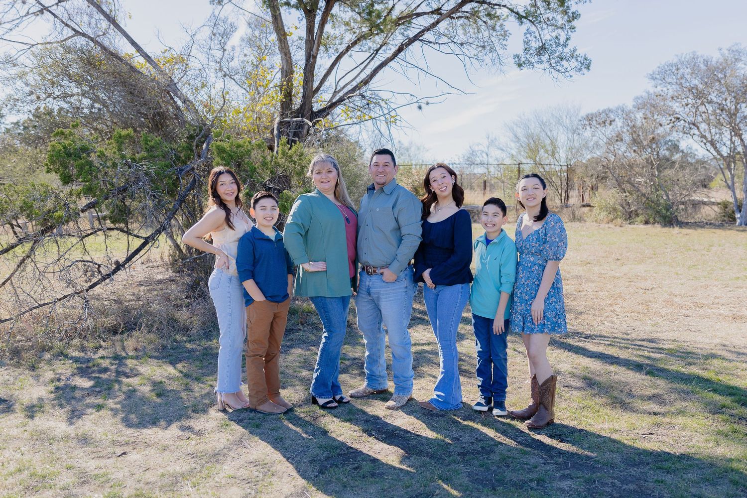 A group of people in casual wear standing together outdoors on a sunny day with trees in the background.