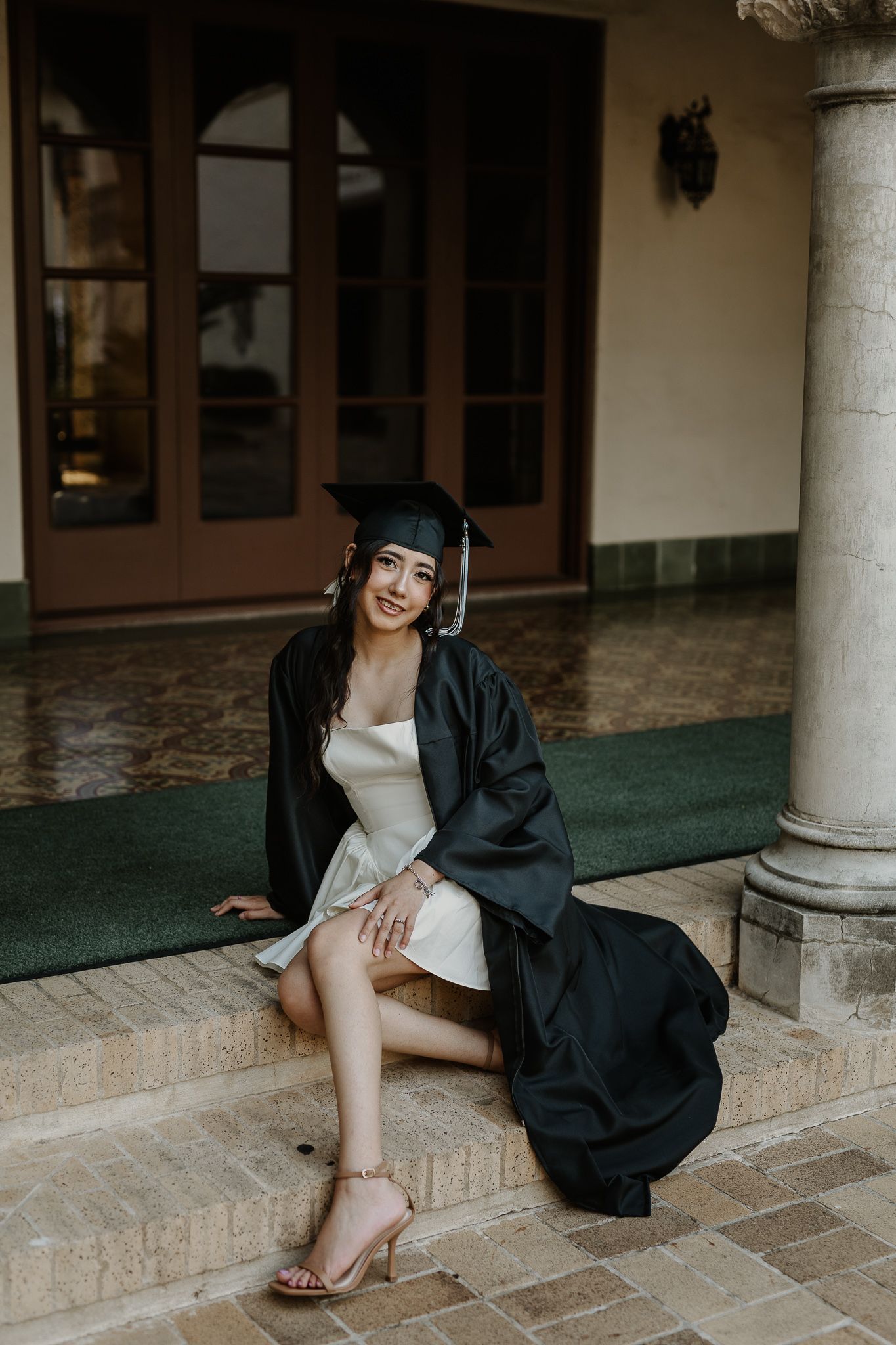 Graduate in traditional black cap and gown sitting on stone steps.